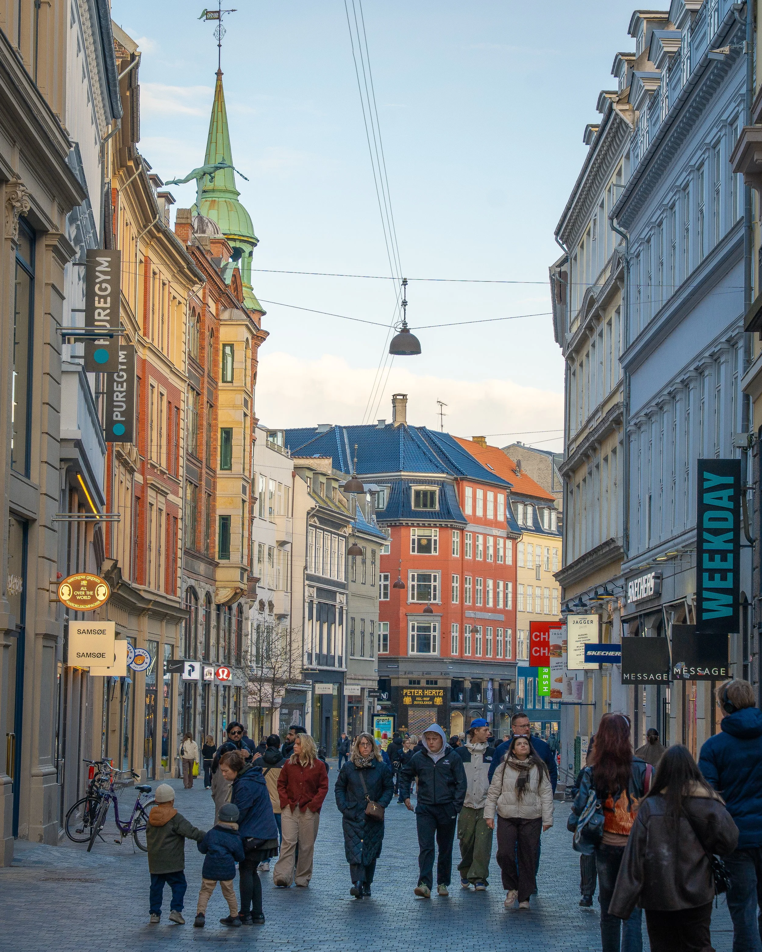 Crowd of people walking down a busy city street lined with shops and tall buildings with colonial architecture, signs, and overhead streetlights, during daytime.