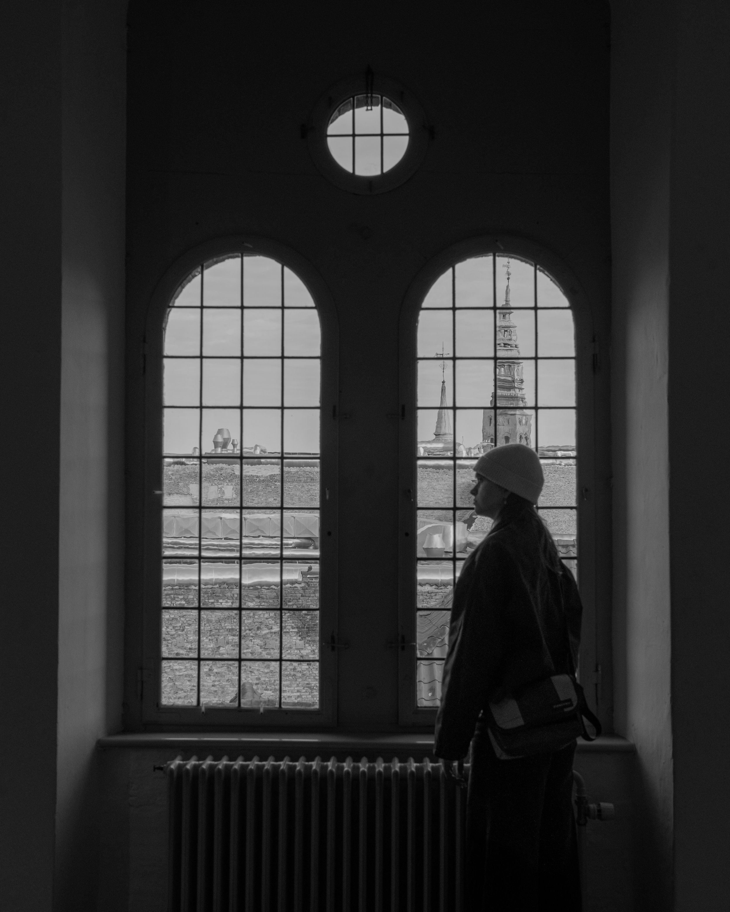 Silhouette of a person wearing a hat and a backpack standing near a large window, with a view of old rooftops and church steeples outside.