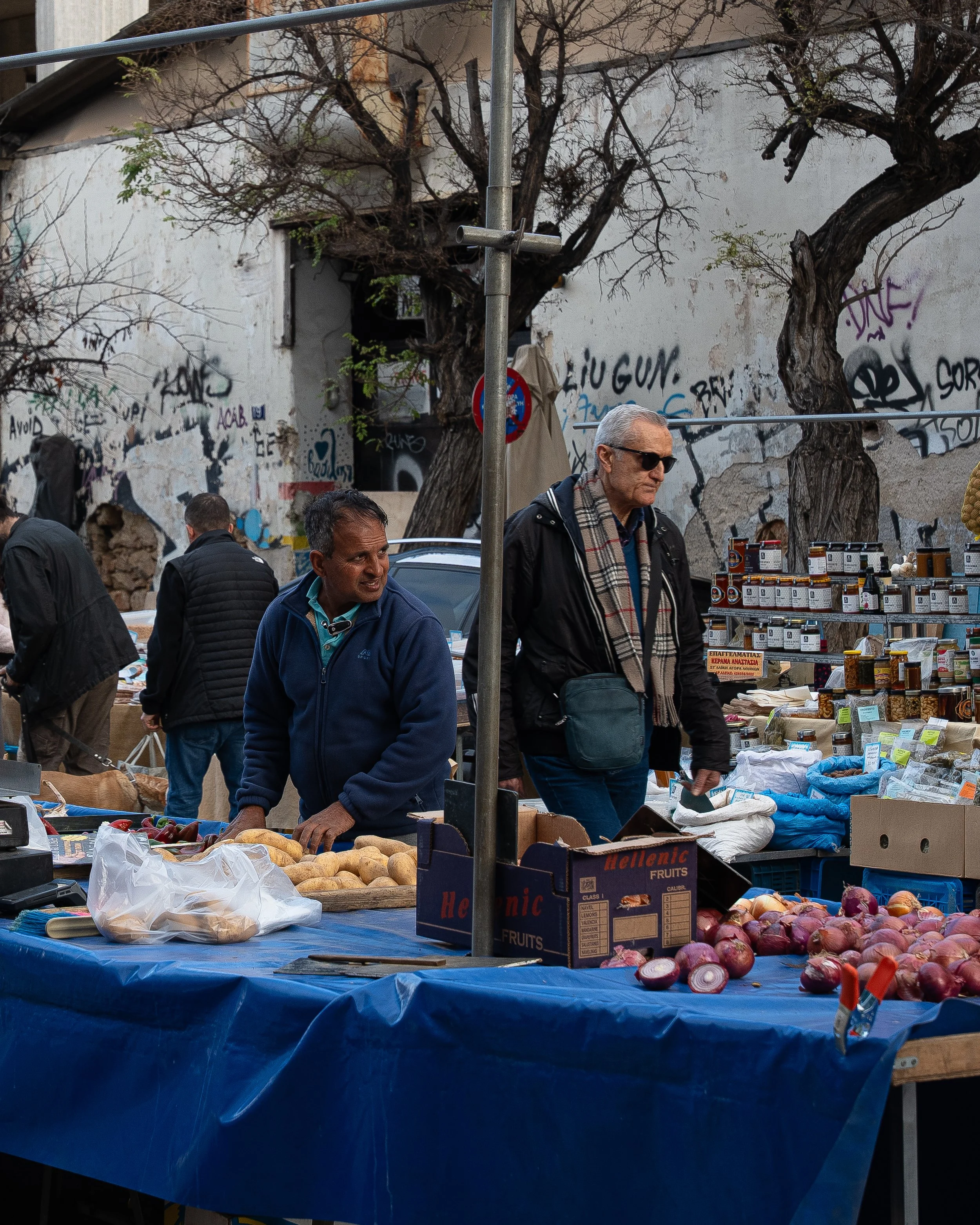 Outdoor market scene with people browsing produce, including onions and root vegetables, under a blue tarp. Graffiti and graffiti-covered wall with trees in the background.