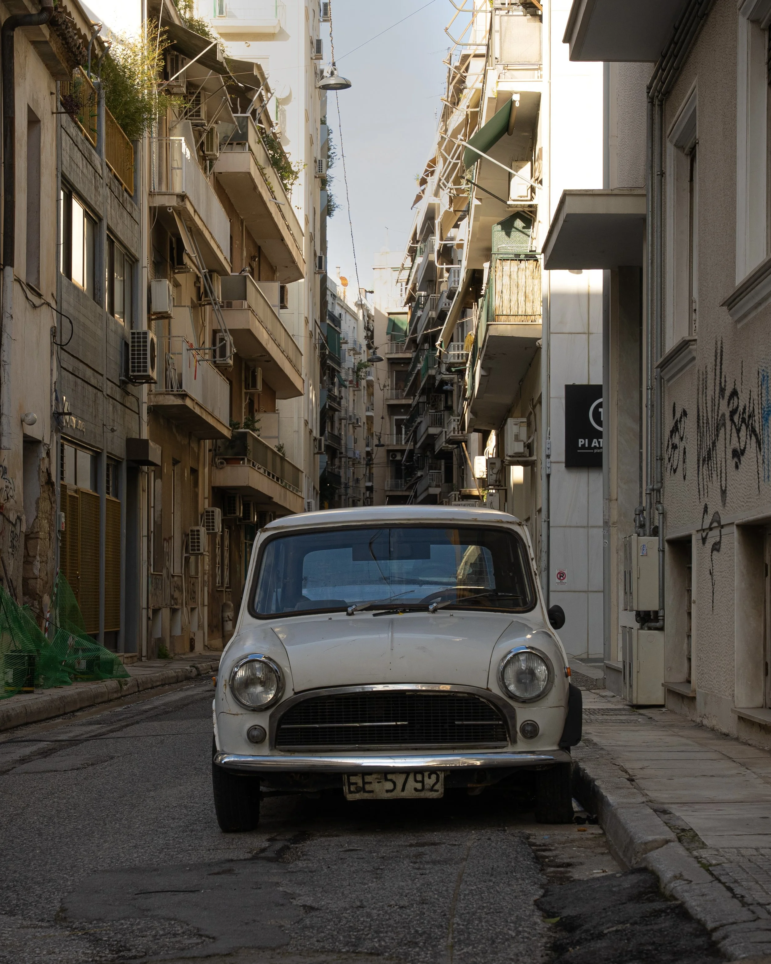 A vintage white Fiat 500 car parked on a narrow urban street with apartment buildings on either side. The buildings have balconies with plants and air conditioning units. The street appears slightly worn with cracks and patches on the pavement.