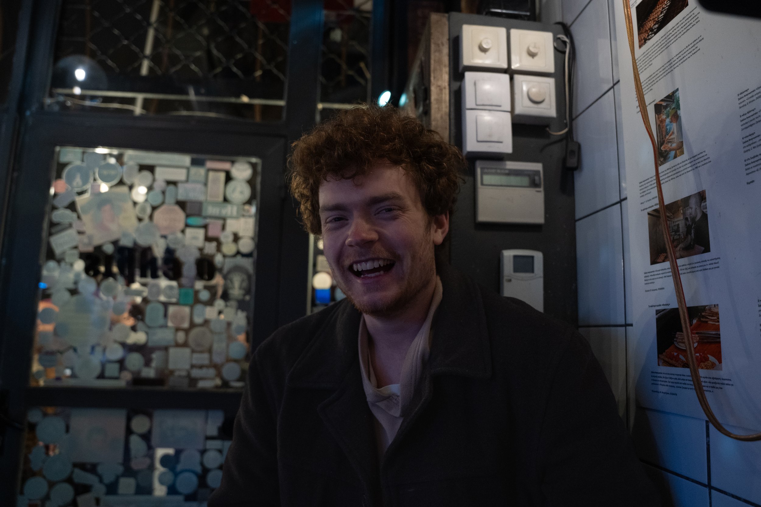 A smiling man with curly hair in a dimly lit environment, with a patterned wall behind him and various objects on the wall to his right.