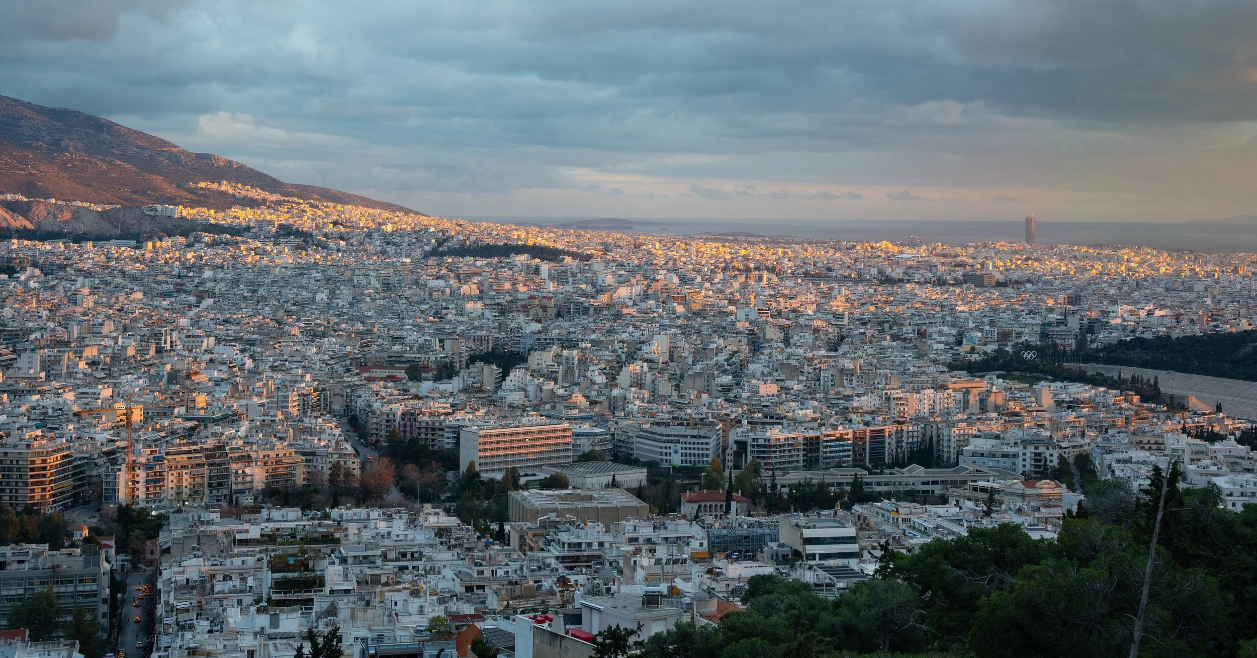Cityscape view of Athens, Greece, with many white buildings, a mountain range on the left, and the sea in the distance under a cloudy sky at sunset.