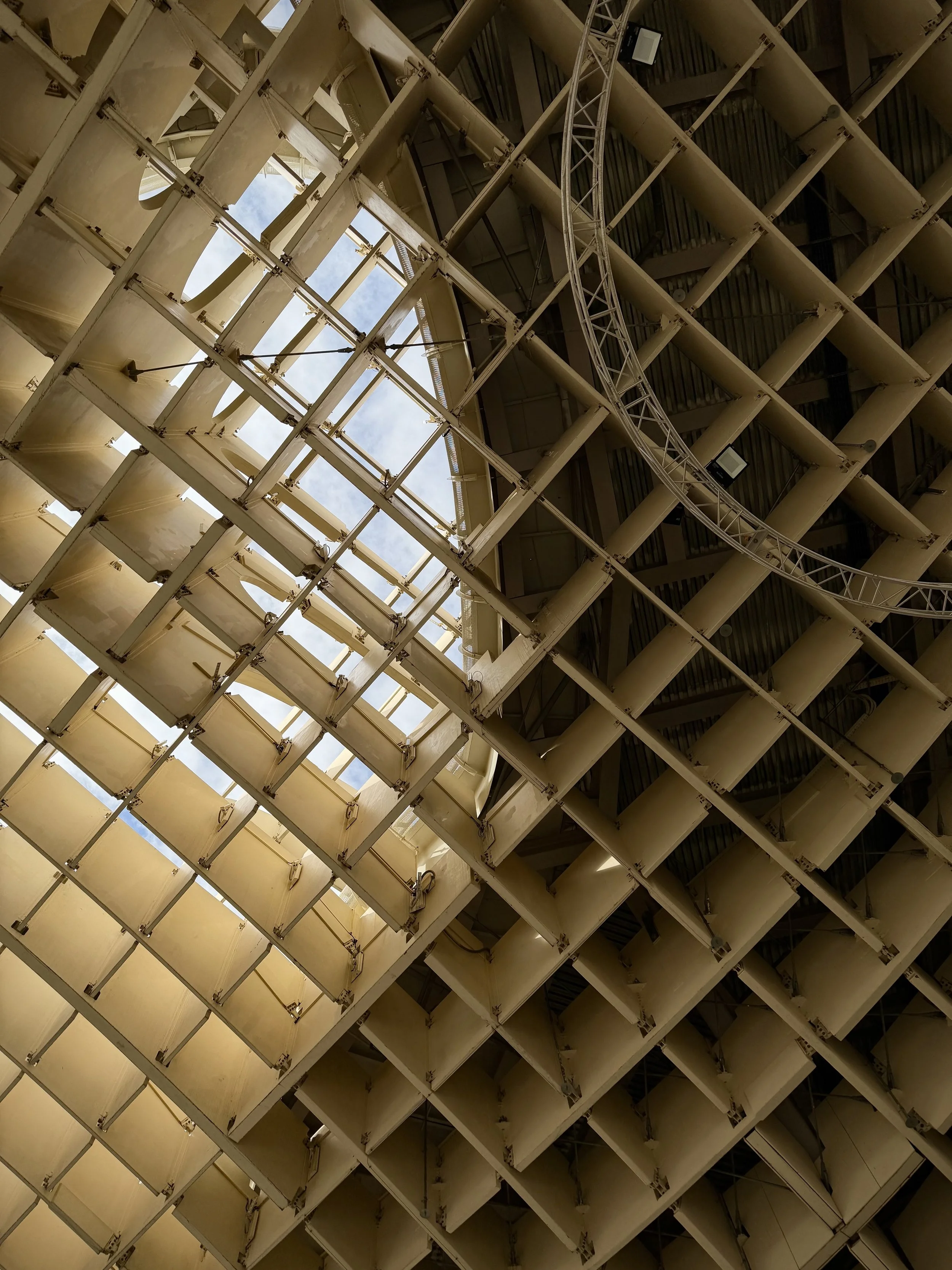Looking up at the underside of a large, modern architectural structure with a grid-like pattern of beige panels and metal beams, and a view of the sky through openings.
