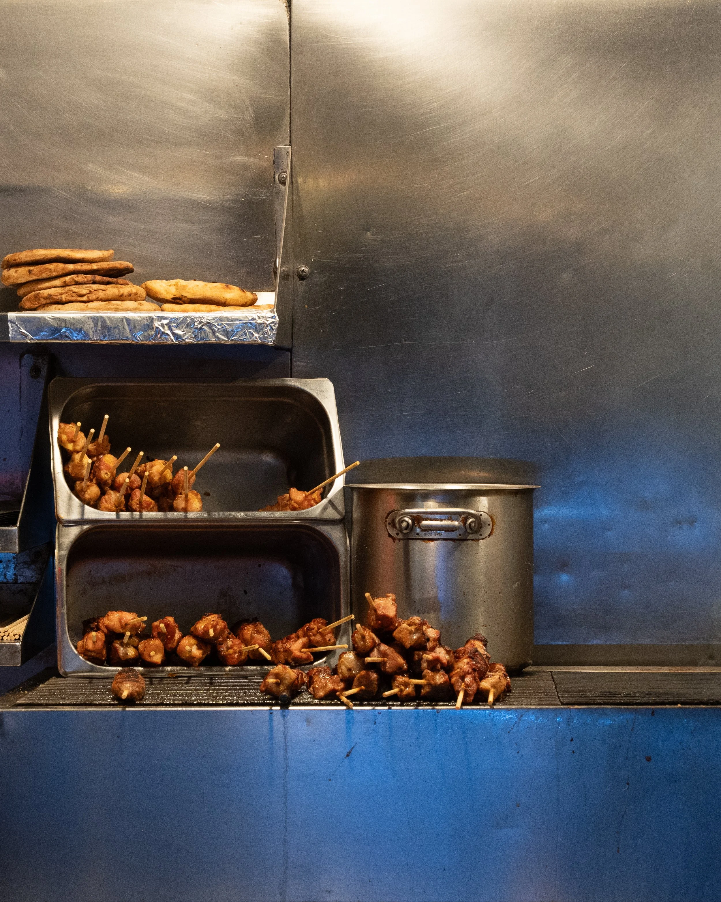 Stacks of grilled skewered meat and chicken in a food stall with a metallic background.