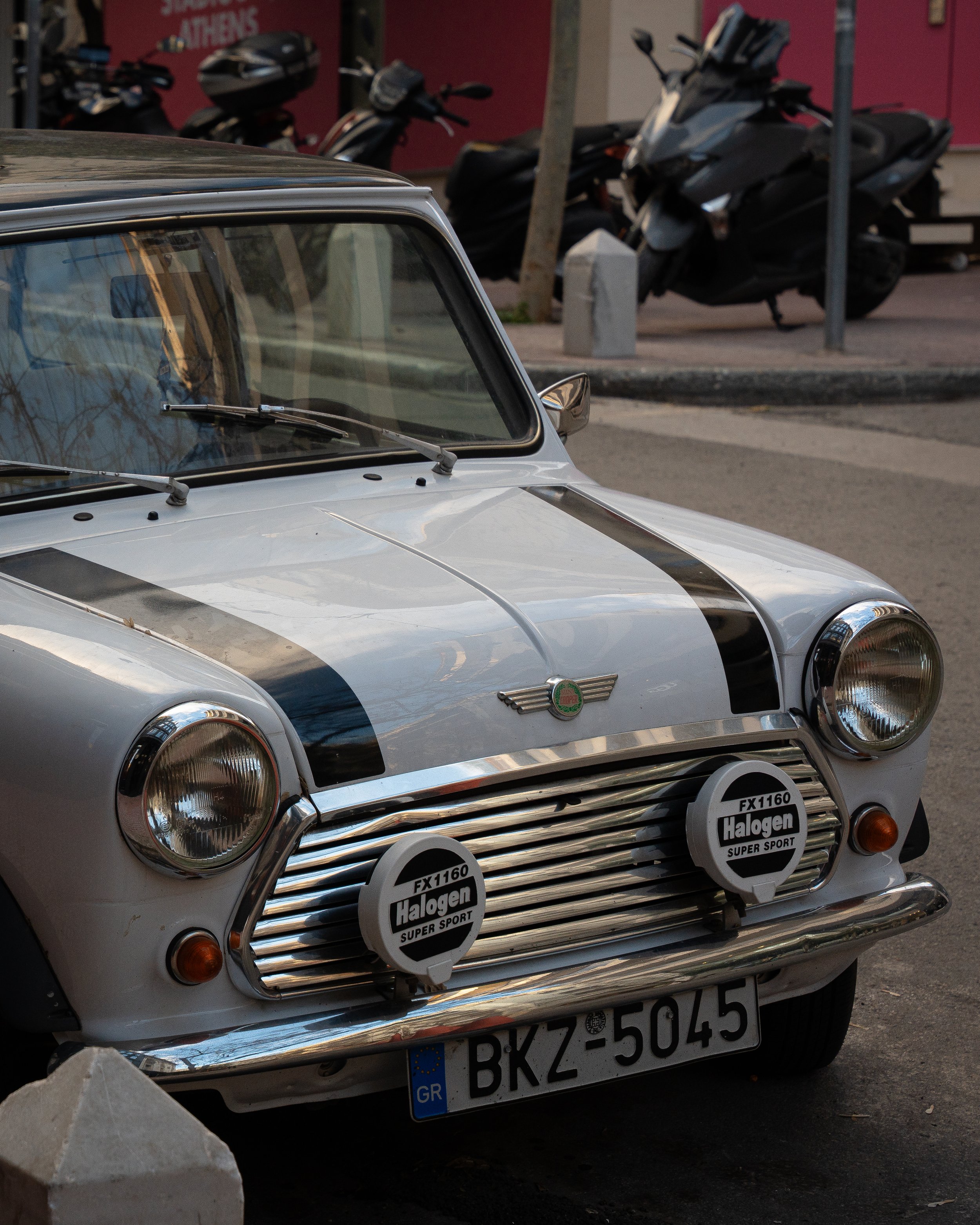 Close-up of a silver vintage car with a white front bumper and black stripe details, parked on the street in front of two black motorcycles. The car has a badge on the front that reads 'Halogen Super Sport' and a European Union license plate from Gre
