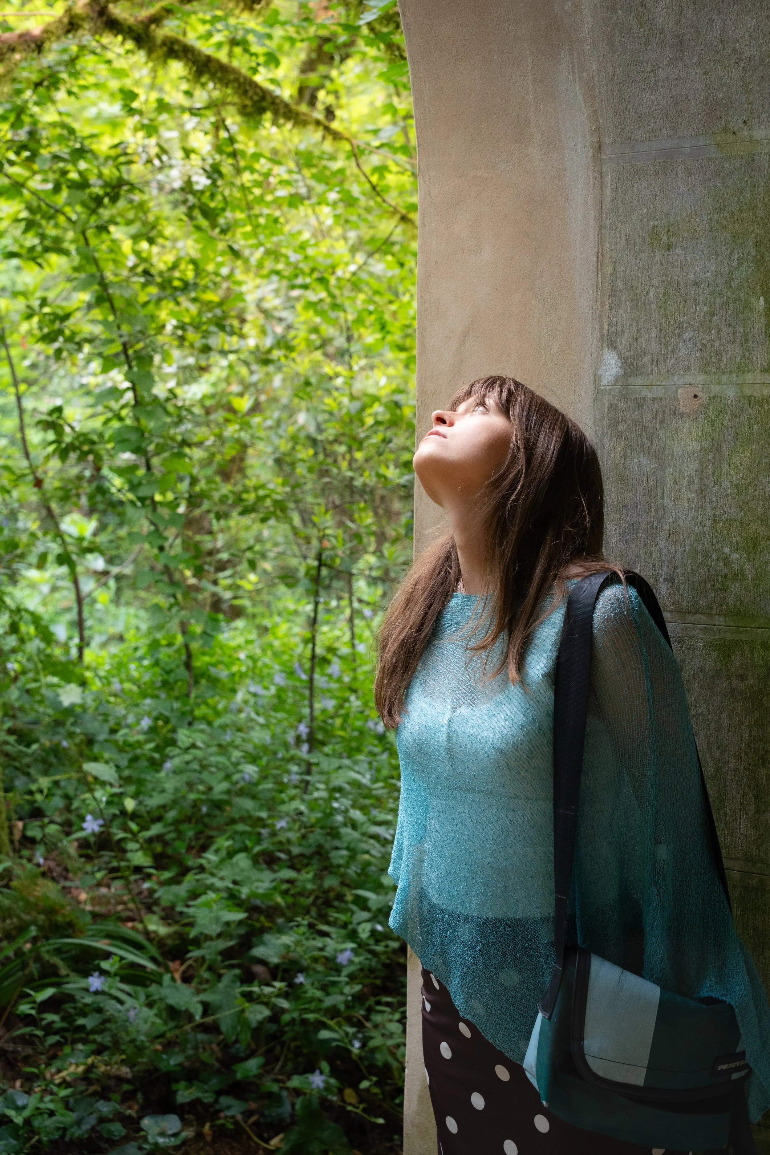 A woman with long brown hair wearing a light blue sweater and black pants with white polka dots stands outdoors beside a stone or concrete pillar. She is looking upwards with a thoughtful expression at the lush green foliage and trees in the backgrou