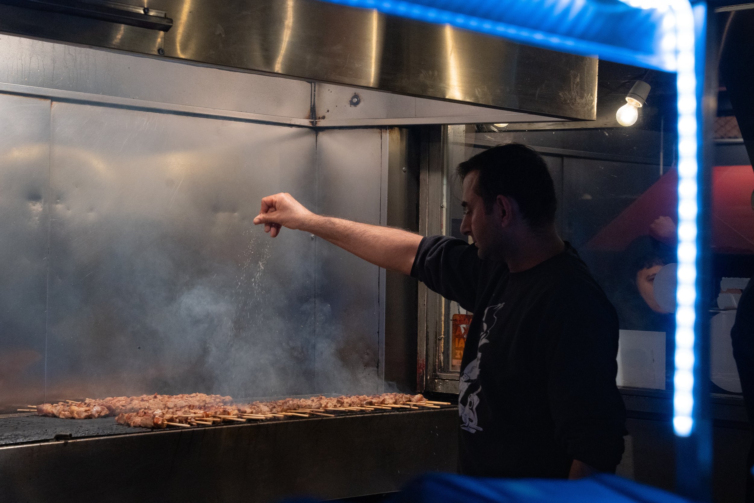 A man grilling skewers of meat in a commercial kitchen or restaurant setting, with a metal grill, a wall, and a bright light in the background.