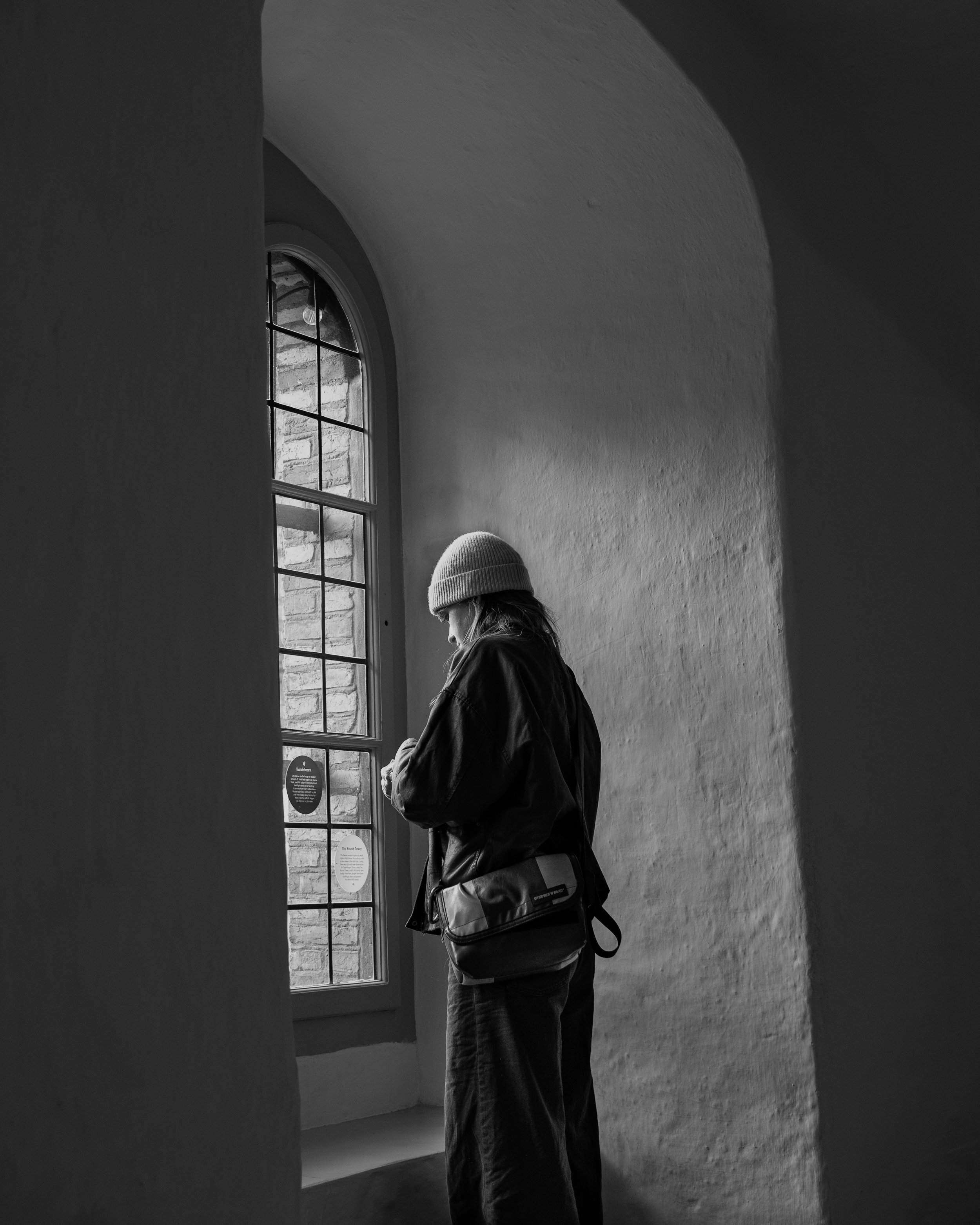 A person standing near a large window with a wire grid pattern, wearing a beanie, jacket, and carrying a bag, in a room with a curved ceiling and brick wall visible outside the window.