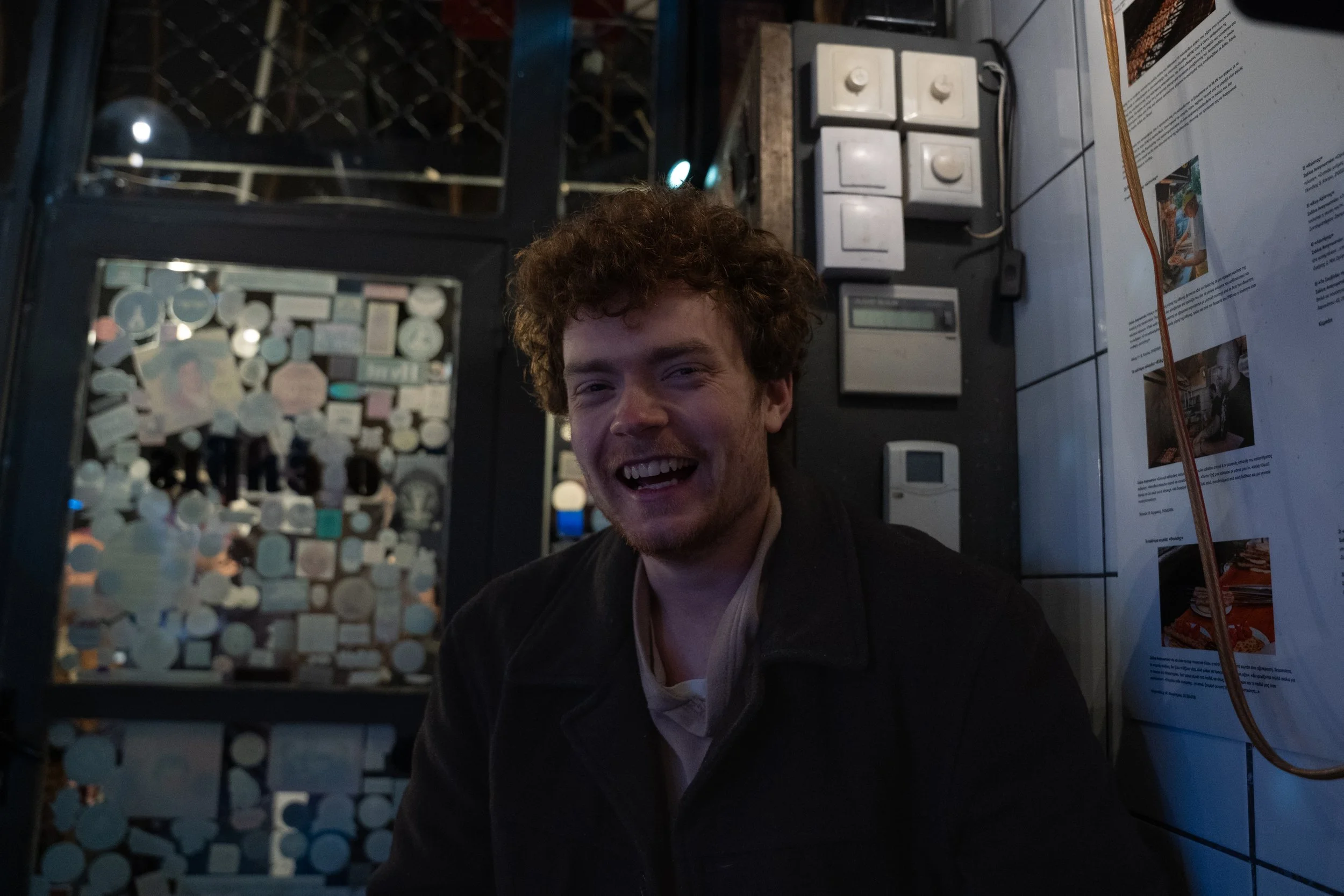 A smiling young man with curly hair sitting next to a glass display case filled with coins, in what appears to be a museum or exhibit setting.
