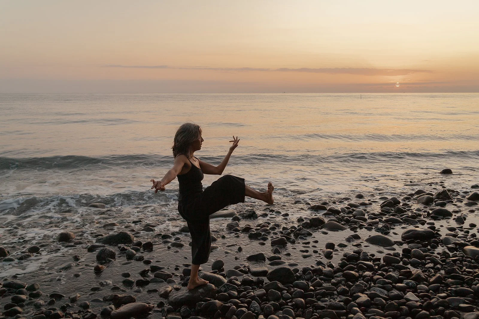A woman practicing yoga on a rocky beach at sunrise or sunset, standing on one leg with the other leg raised, and arms outstretched.