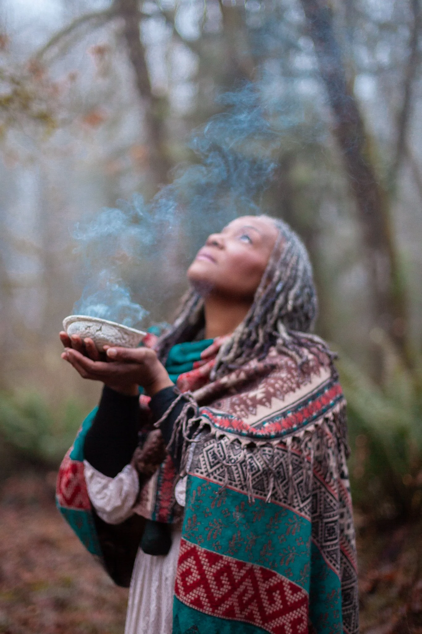 A woman with braided hair and colorful patterned shawl holding a bowl with smoke or steam in a forest setting.