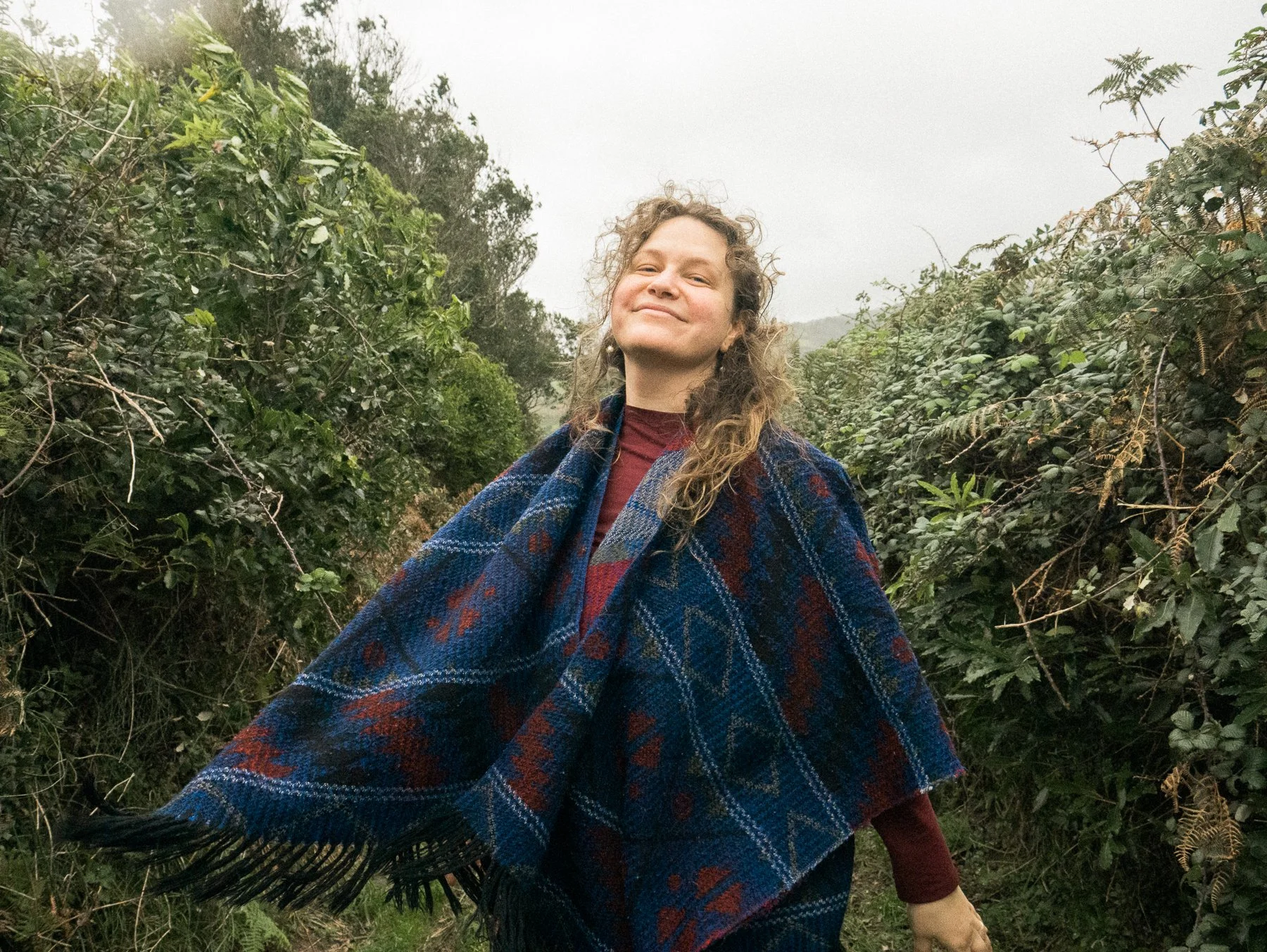 A woman standing outdoors in a lush, green, bush-filled environment, smiling with a confident expression, wearing a colorful plaid shawl.