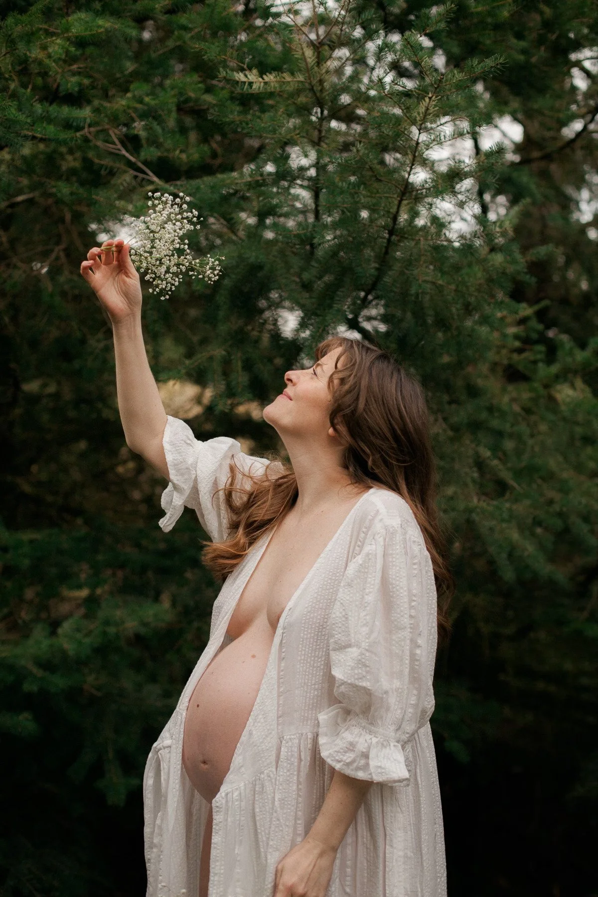 A pregnant woman in a white dress standing outdoors among green trees, holding a small bouquet of white flowers and looking up at the flowers with a smile.