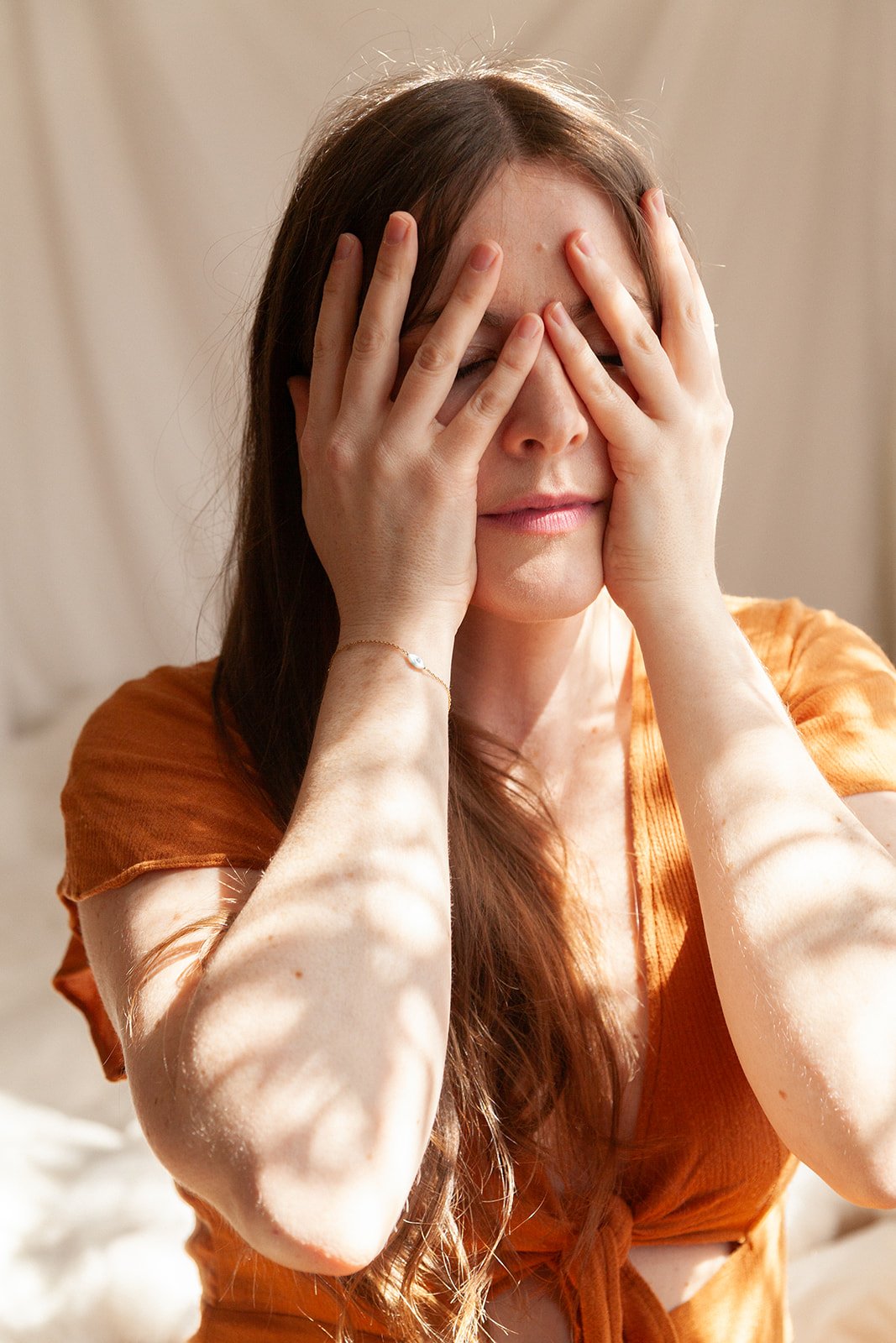 A woman with long brown hair covering her face with her hands, wearing an orange top and a delicate bracelet, in a softly lit room.
