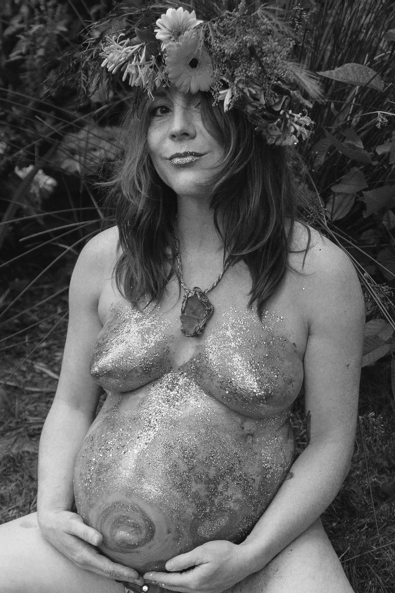 A woman with a flower crown on her head, glitter on her lips and body, and a large necklace, holding her pregnant belly, outdoors surrounded by plants.