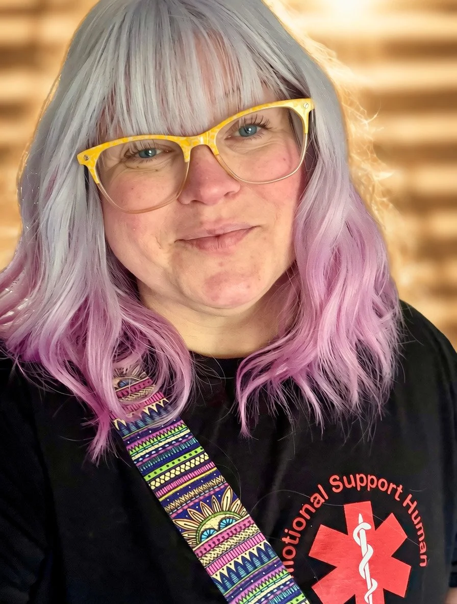 A woman with gray and pastel purple hair, wearing yellow glasses and a black shirt with a red medical support symbol with red font stating emotional support human, standing in front of a window with the sun shining through wooden blinds.