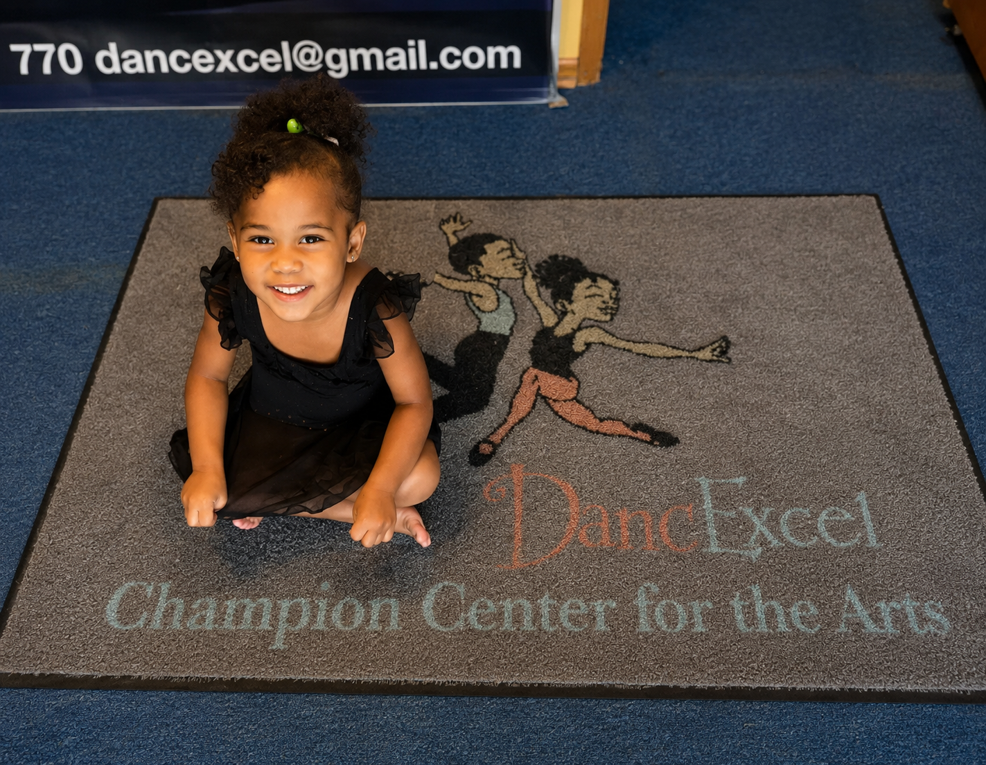 A young girl with curly hair tied back, wearing a black dress, is kneeling on a gray carpet with a dance-themed logo and text. She is smiling at the camera.