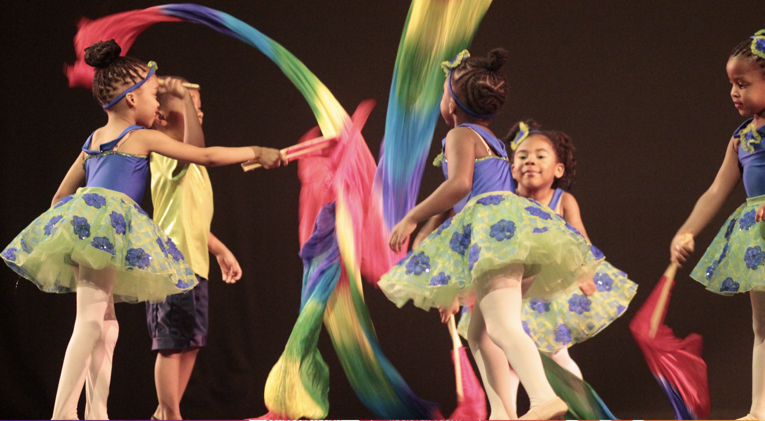 Young children dressed in colorful costumes performing a dance on stage with rainbow ribbons.