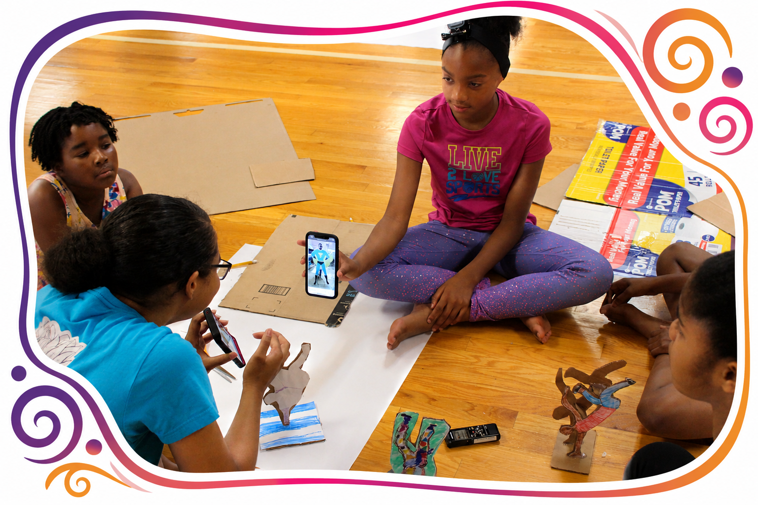 Children sitting on a wooden floor making a project with cardboard and paper, one girl with glasses and two others are taking pictures or recording videos with smartphones.