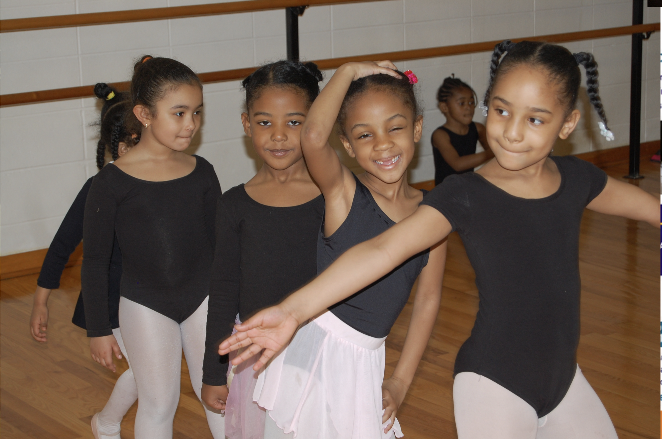 Group of young girls in ballet outfits practicing in a dance studio with wooden floors and ballet barres.