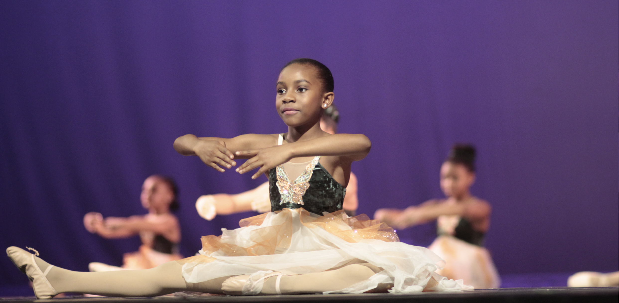 Young girl in ballet costume performing a dance on stage with other dancers in the background.