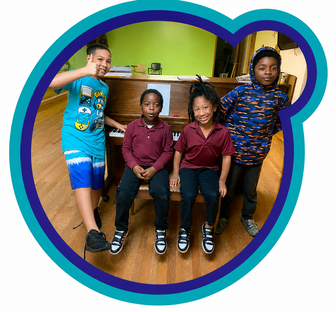 Four children, two boys and two girls, standing and sitting near a piano in a music classroom, all smiling and looking at the camera, with a green wall and classroom furniture in the background.