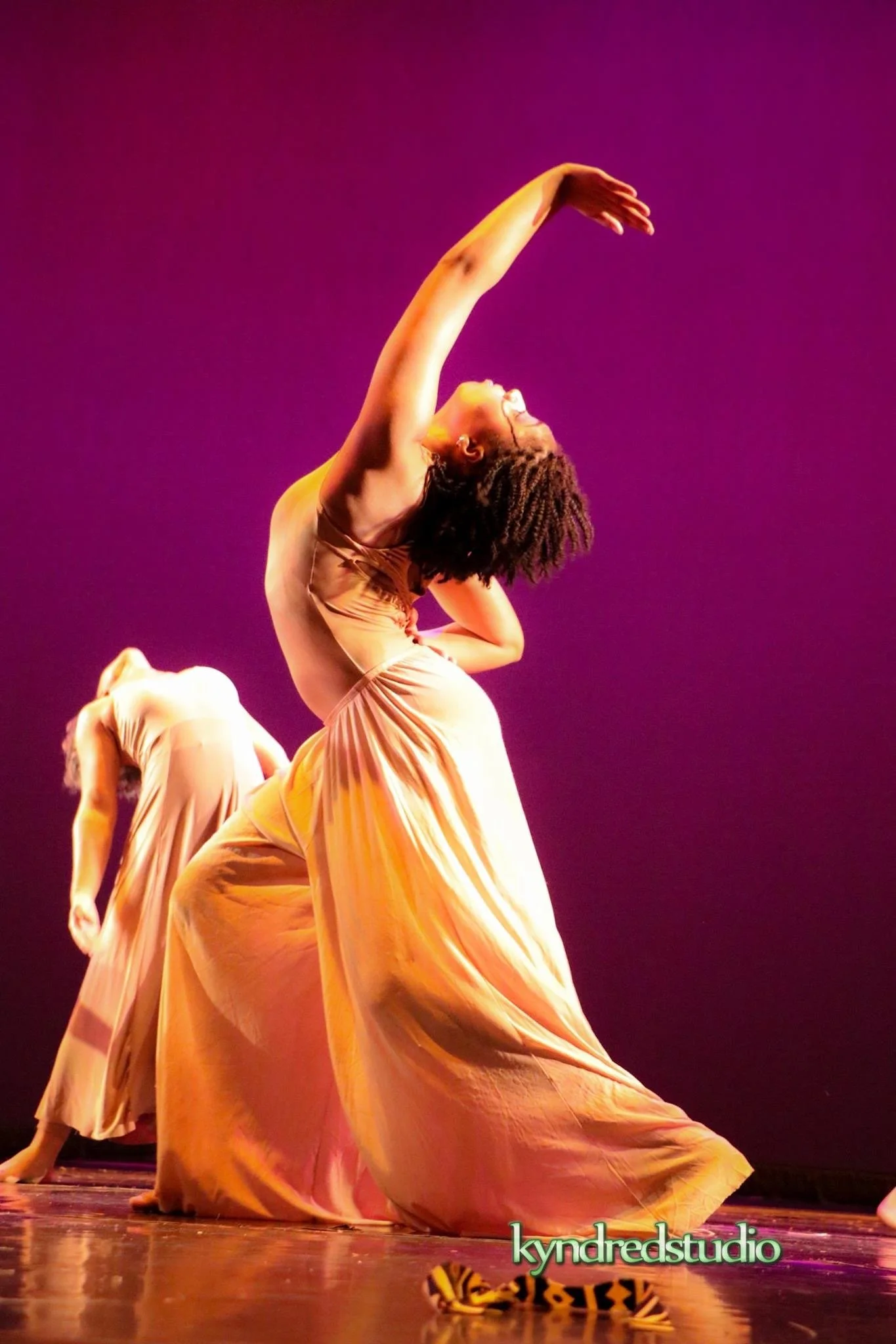 Dancers wearing flowing yellow dresses perform on stage with a purple background, captured from a low angle.