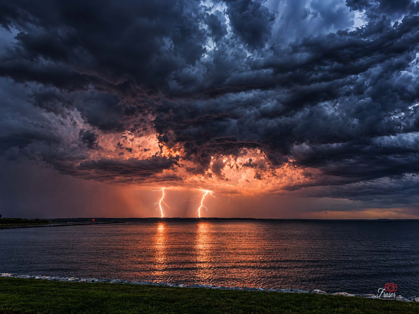 Lightning strikes over the water beneath dramatic storm clouds as an orange glow illuminates the horizon.