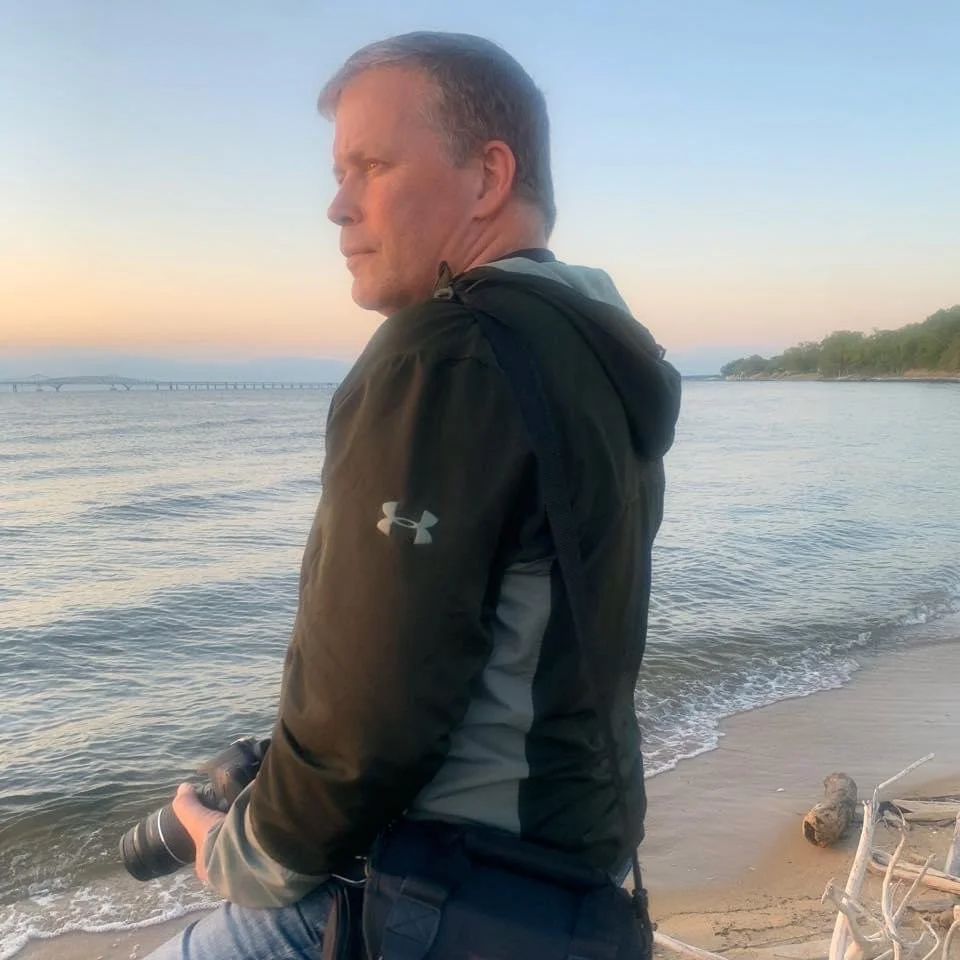 Photographer Christopher Fraser holding his camera on the beach at sunset with calm water and a bridge in the background.