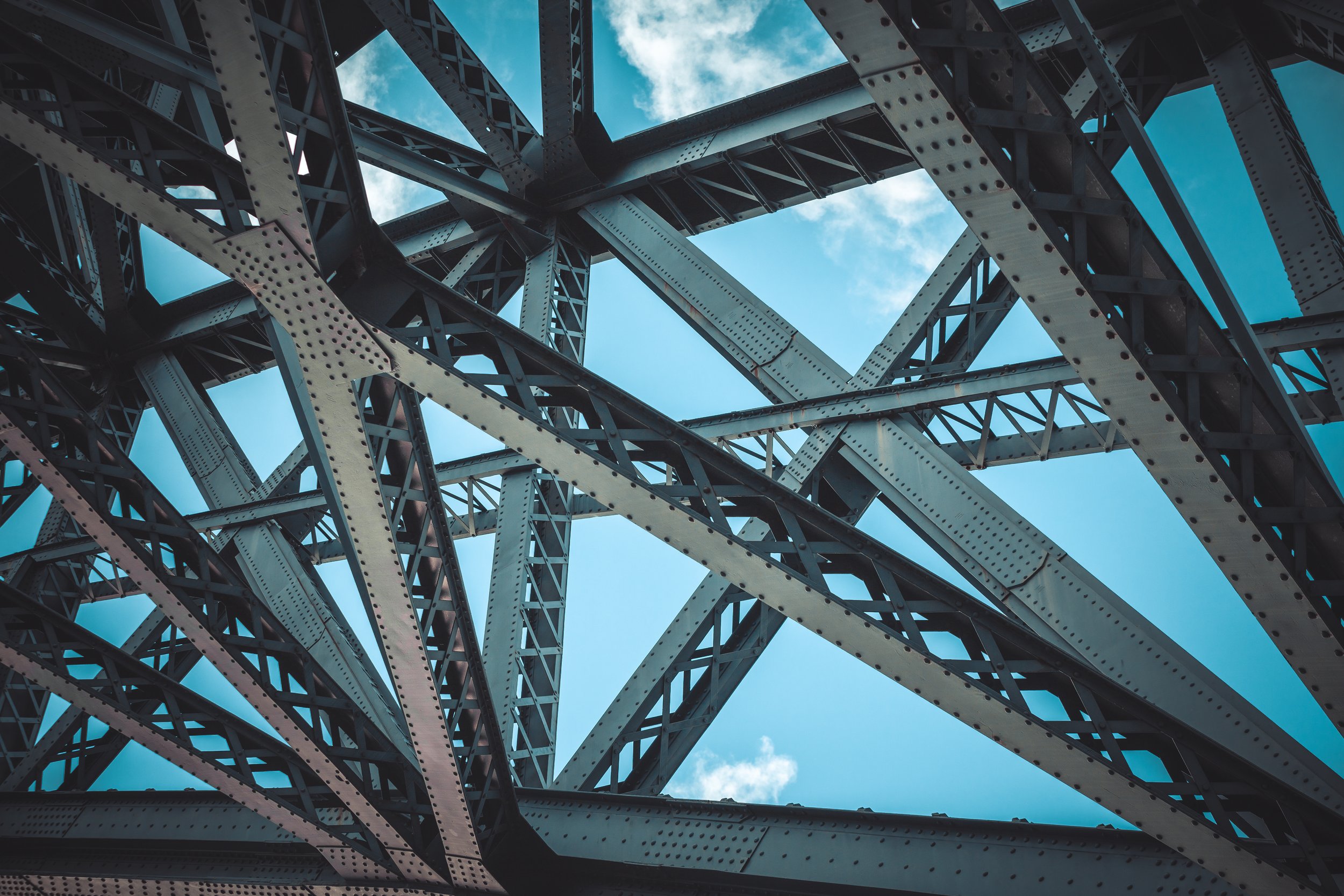 Close-up view of the steel framework beneath a truss bridge, showing intersecting beams and riveted connections against a blue sky.