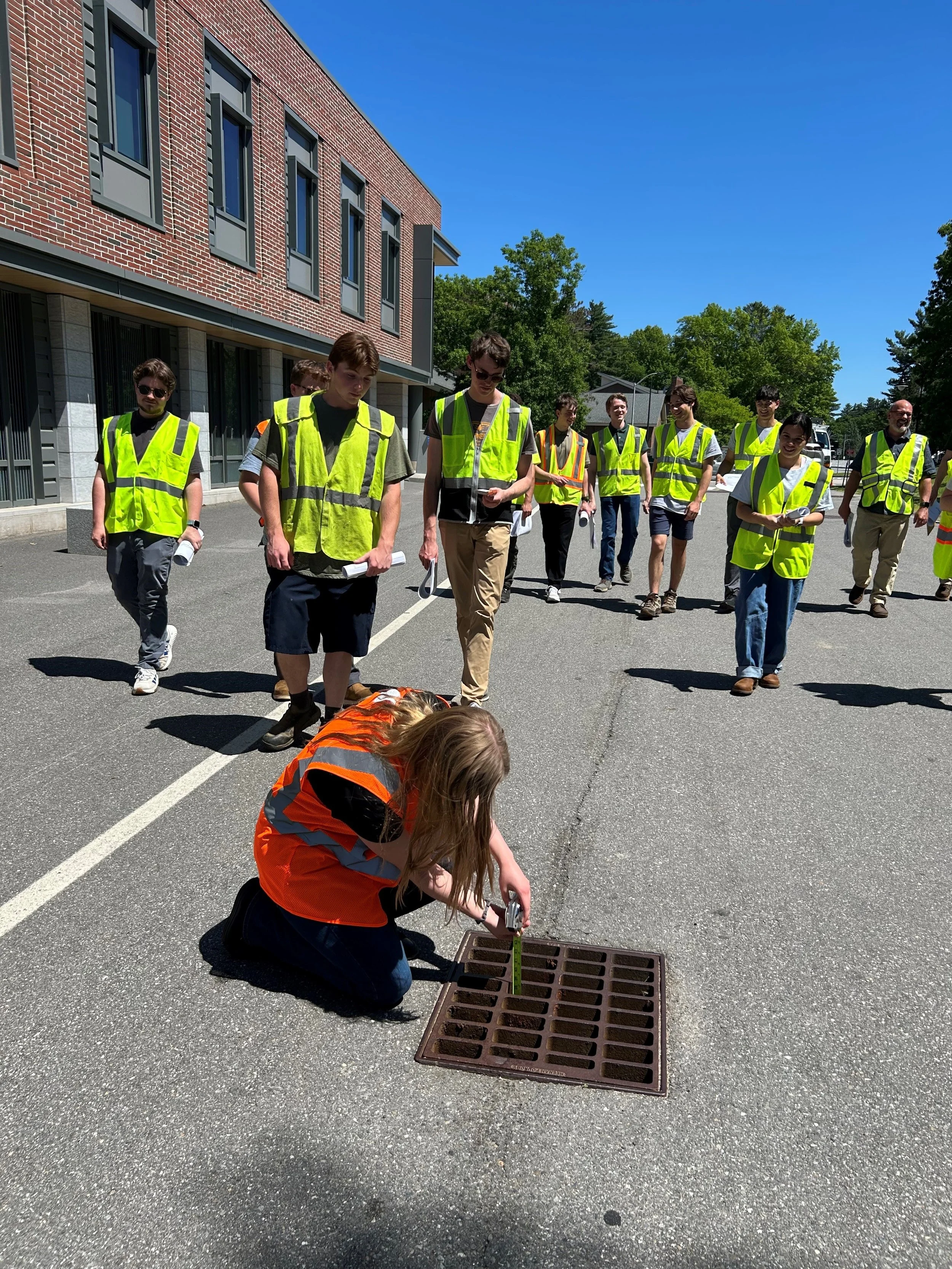 Group of people in high vis yellow vests walk towards someone crouching at a storm drain wearing high vis orange vest. She's measuring something at drain.