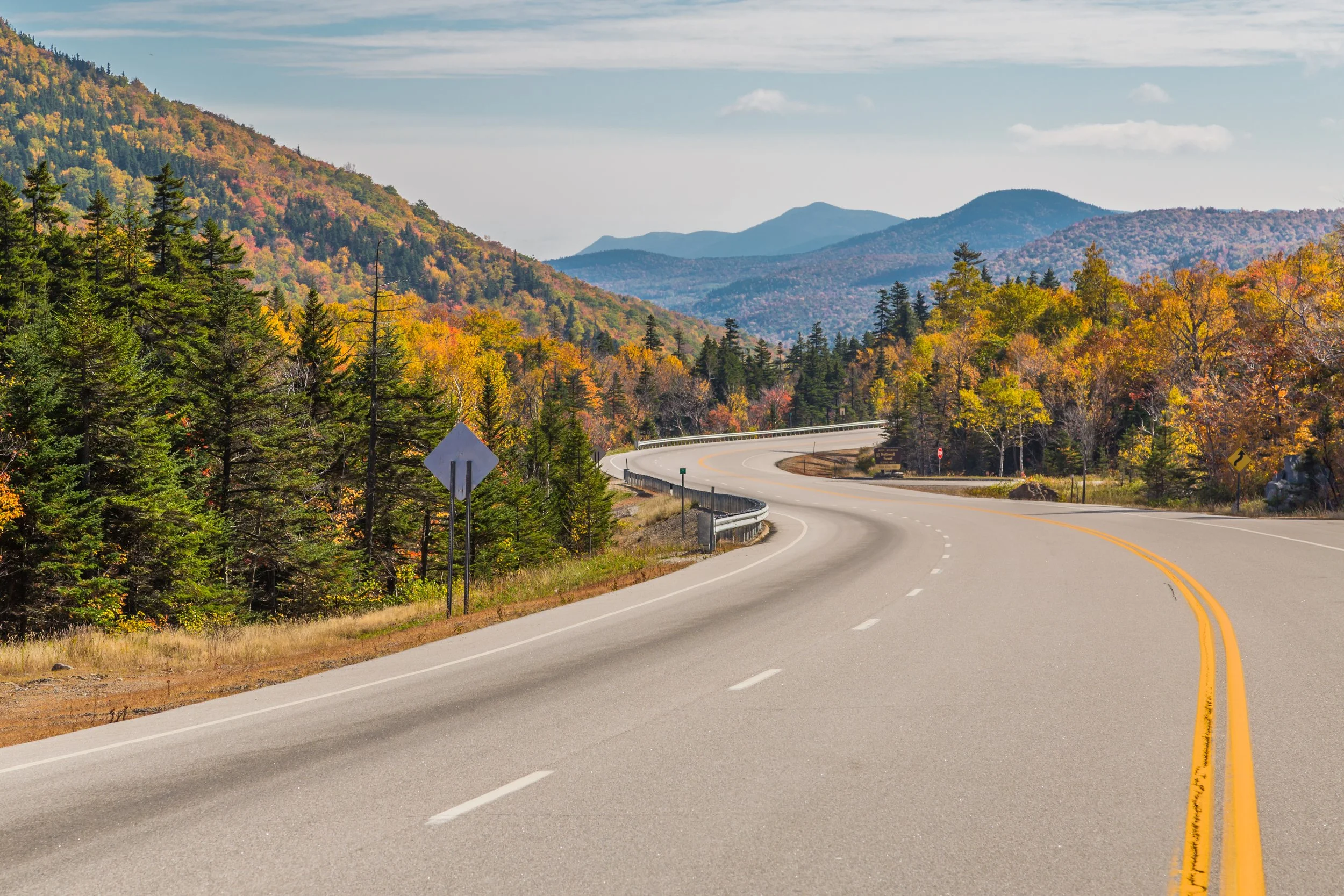 Curving highway winding through a mountainous landscape with colorful autumn foliage and distant peaks.