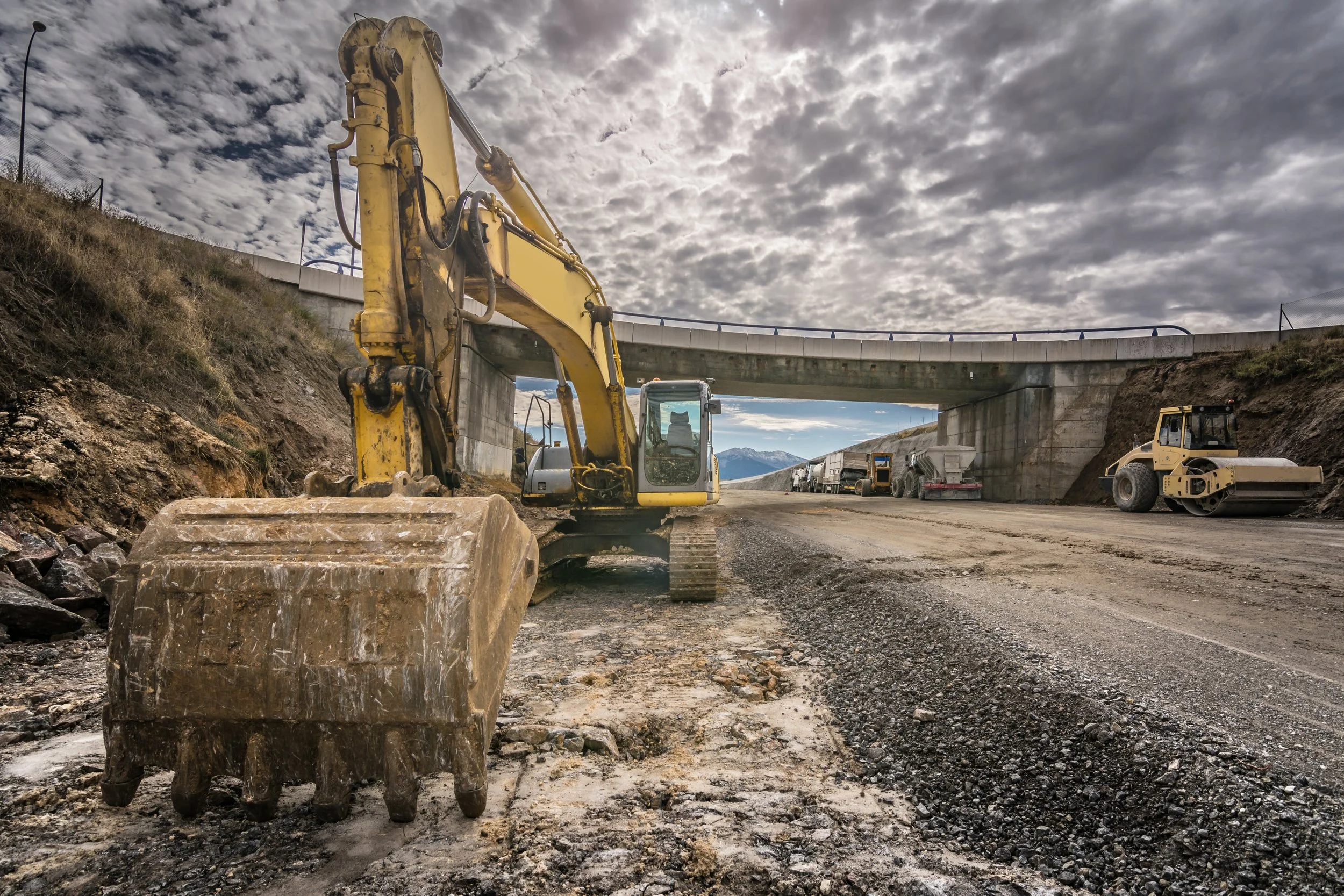 Large yellow excavator parked on a gravel roadway beneath a concrete overpass at an active construction site, with a road roller and construction vehicles visible in the background under a cloudy sky.