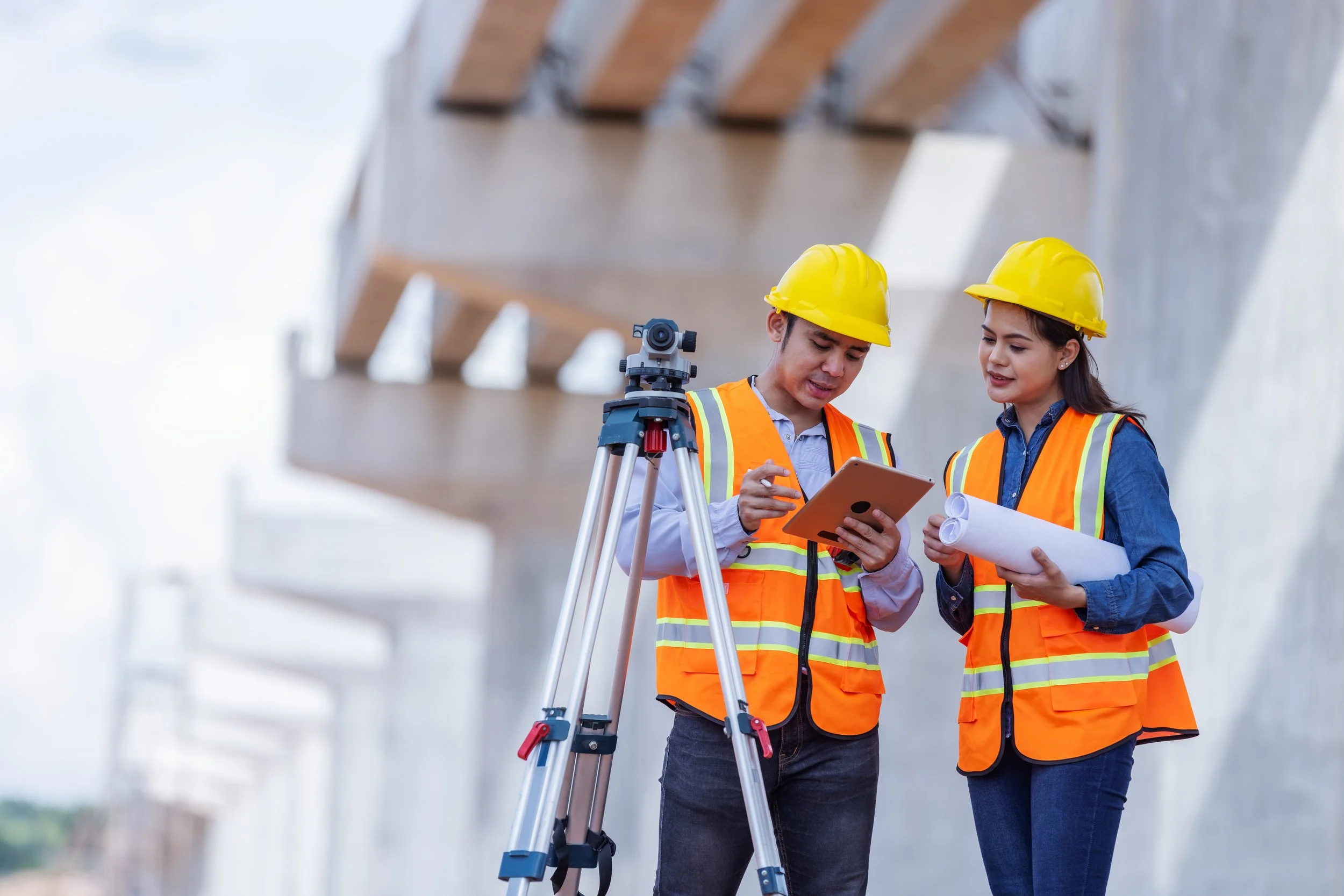 Two construction workers wearing orange safety vests and yellow hard hats review plans on a tablet beside a surveying tripod at a construction site with large concrete structures in the background.