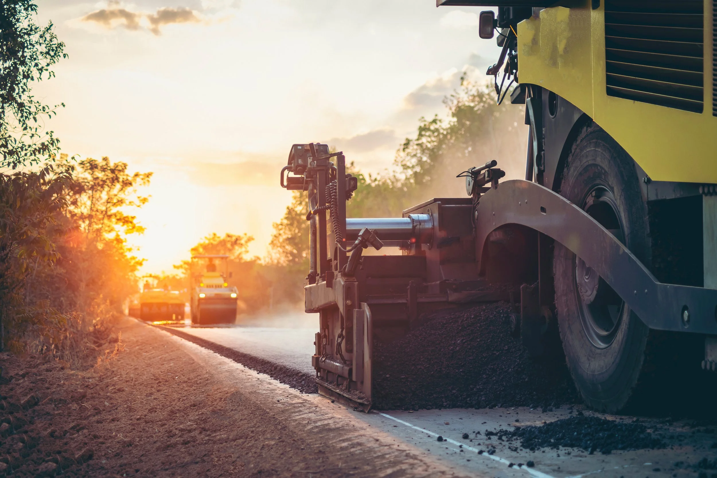 Asphalt paving machine lays new roadway surface while a road roller follows behind during sunset.