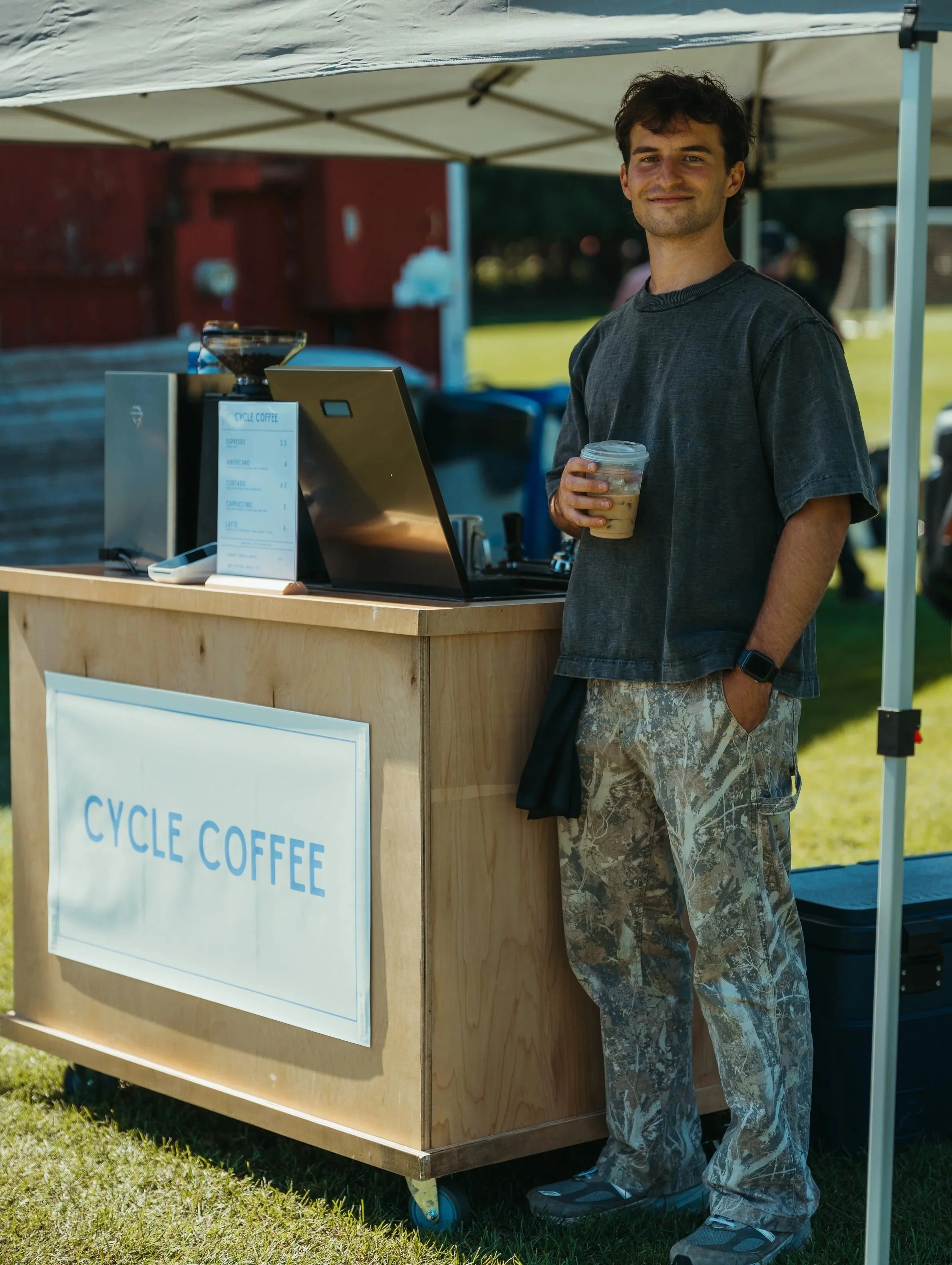 A young man standing at a coffee stand outdoors, holding a cold coffee drink in a plastic cup, wearing a black t-shirt, camouflage pants, and sneakers, under a white canopy with a menu sign that says 'Cycle Coffee'.
