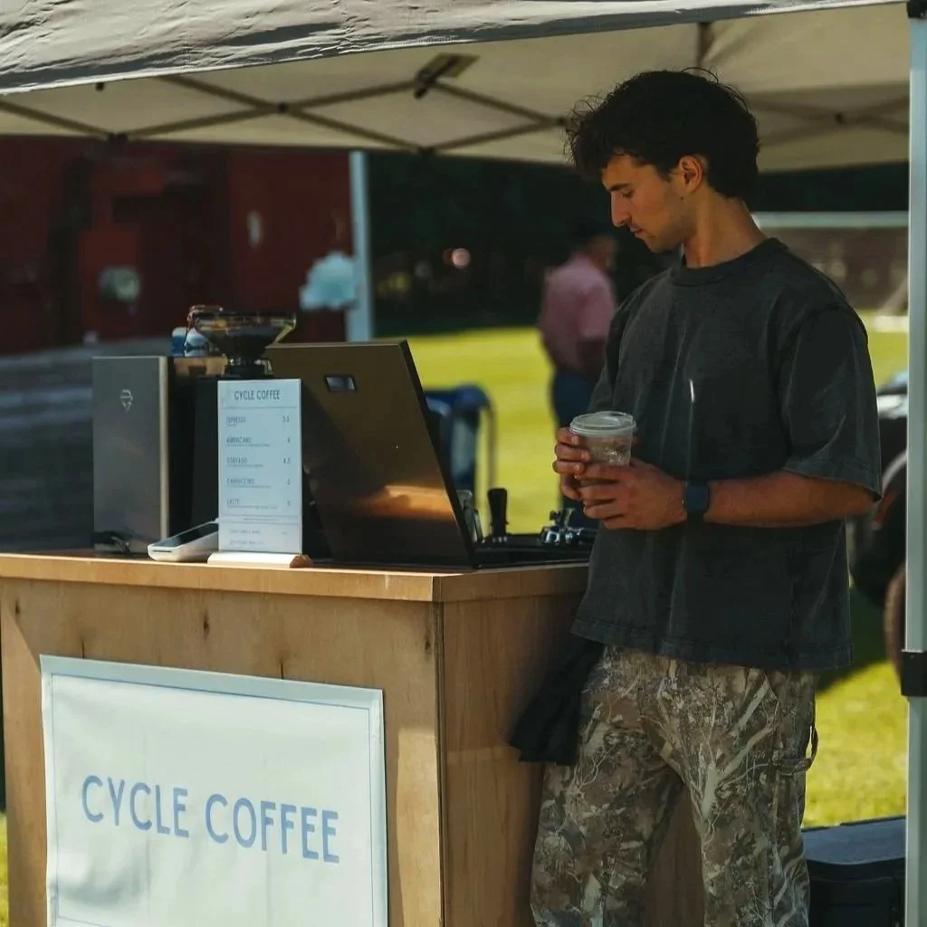 A young man at a coffee stand holding a plastic cup of iced coffee, wearing a black t-shirt and camouflage pants, standing under a beige canopy.