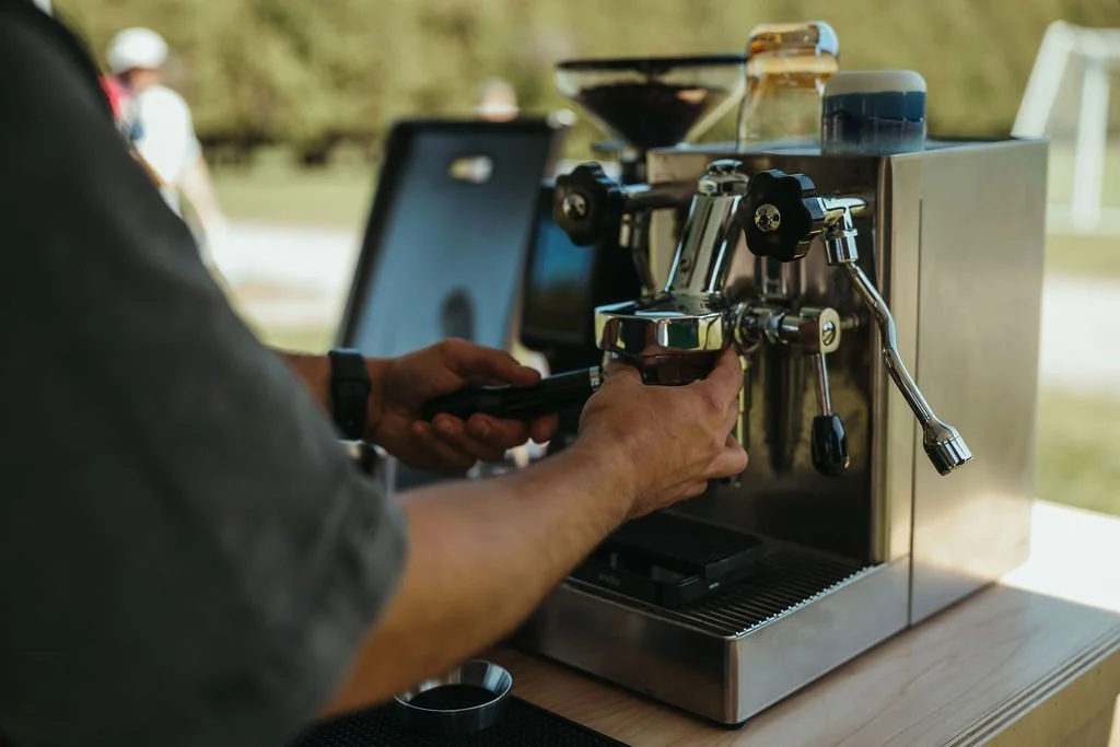 Close-up of a person operating a stainless steel espresso machine outdoors, with a tablet and coffee cups nearby.