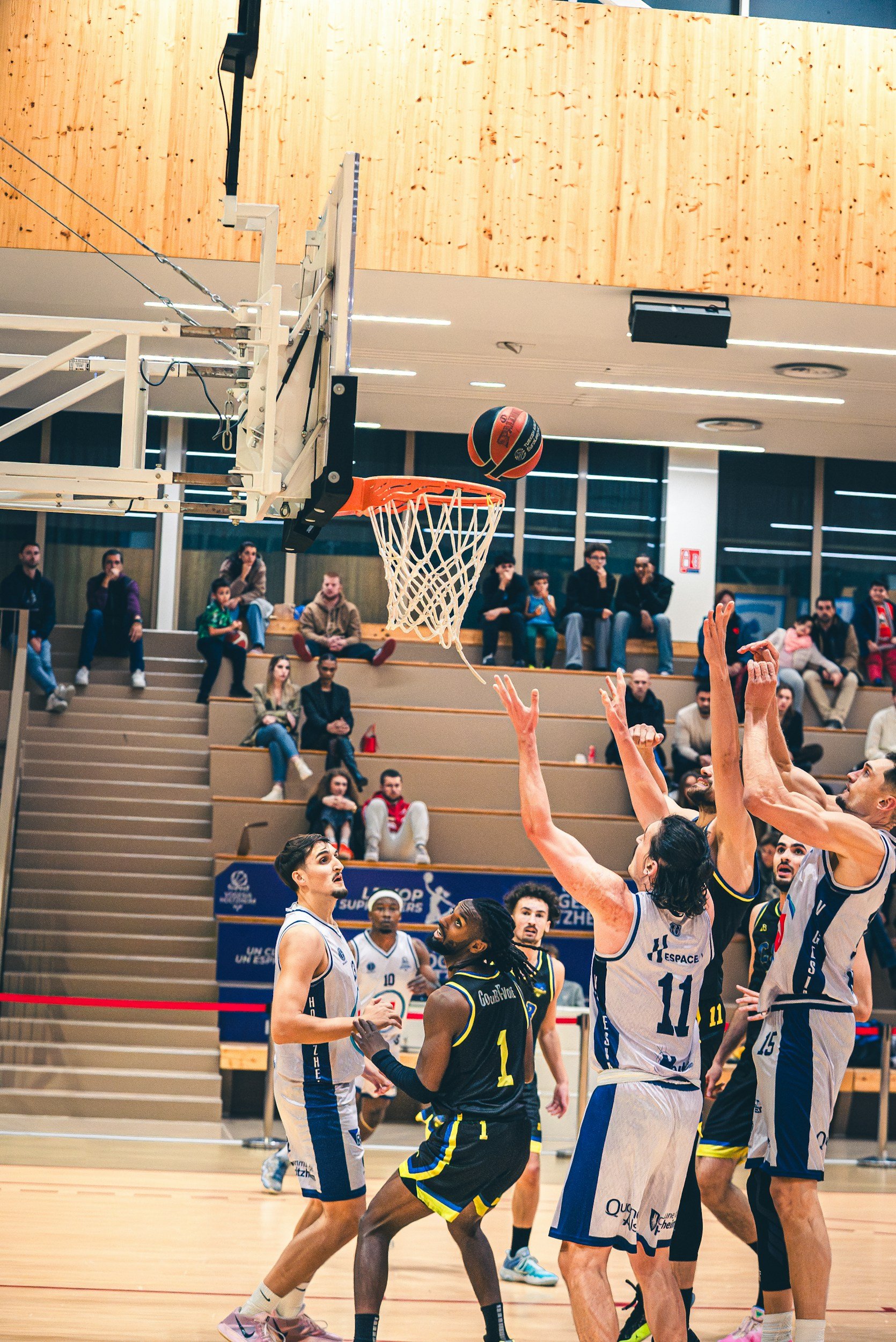 Basketball game in progress with players reaching for the ball near the basket, spectators watching from the bleachers.