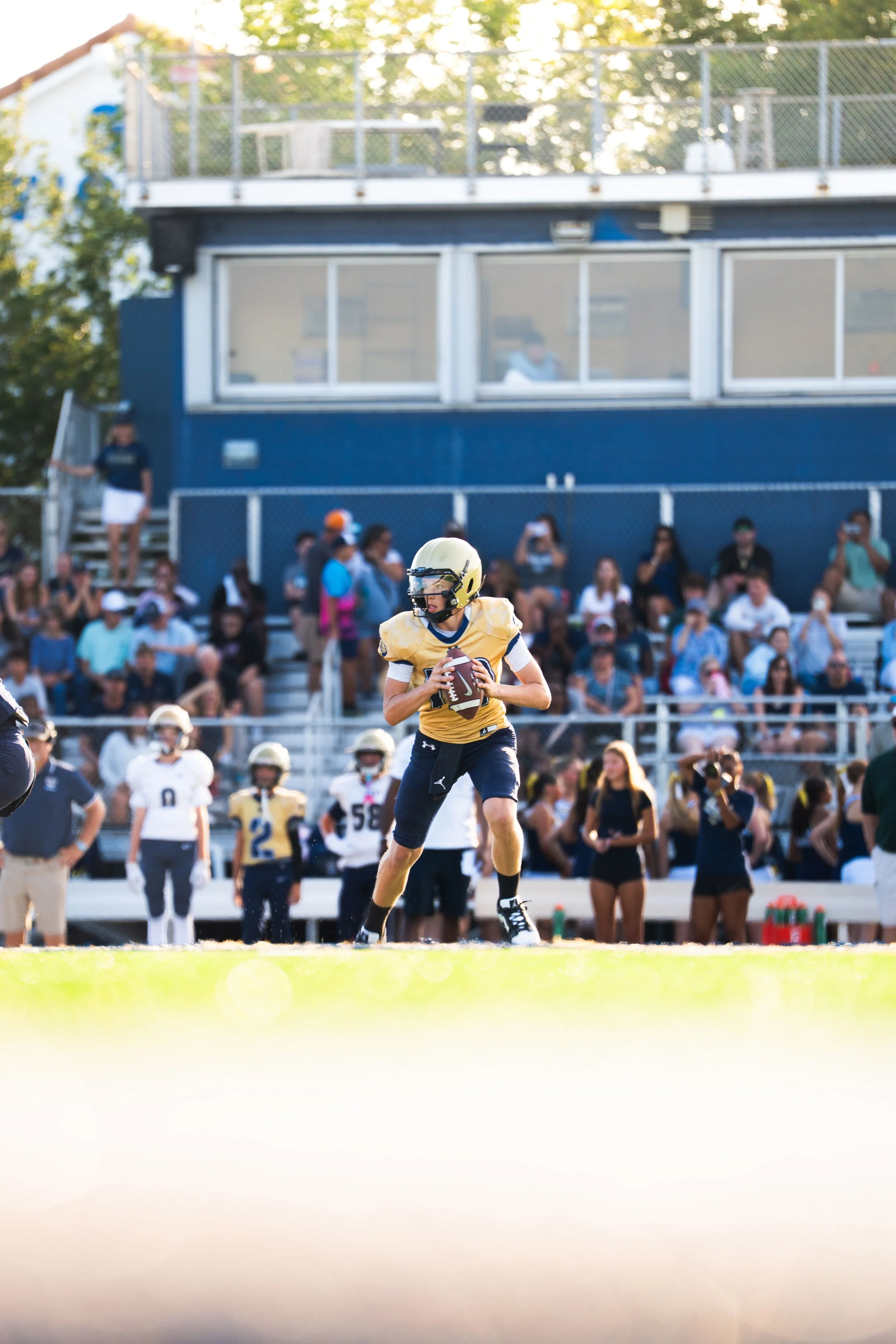 A football player in a yellow jersey and black helmet is standing on the field holding a football, with a crowd and a blue building in the background.