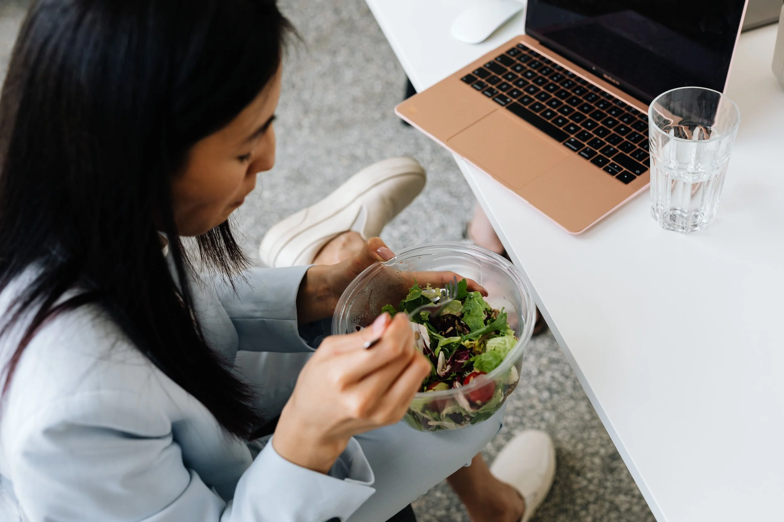 A woman is sitting at a white table, eating a salad from a clear plastic container with a fork. On the table, there is a pink laptop, a white computer mouse, and a clear glass of water.
