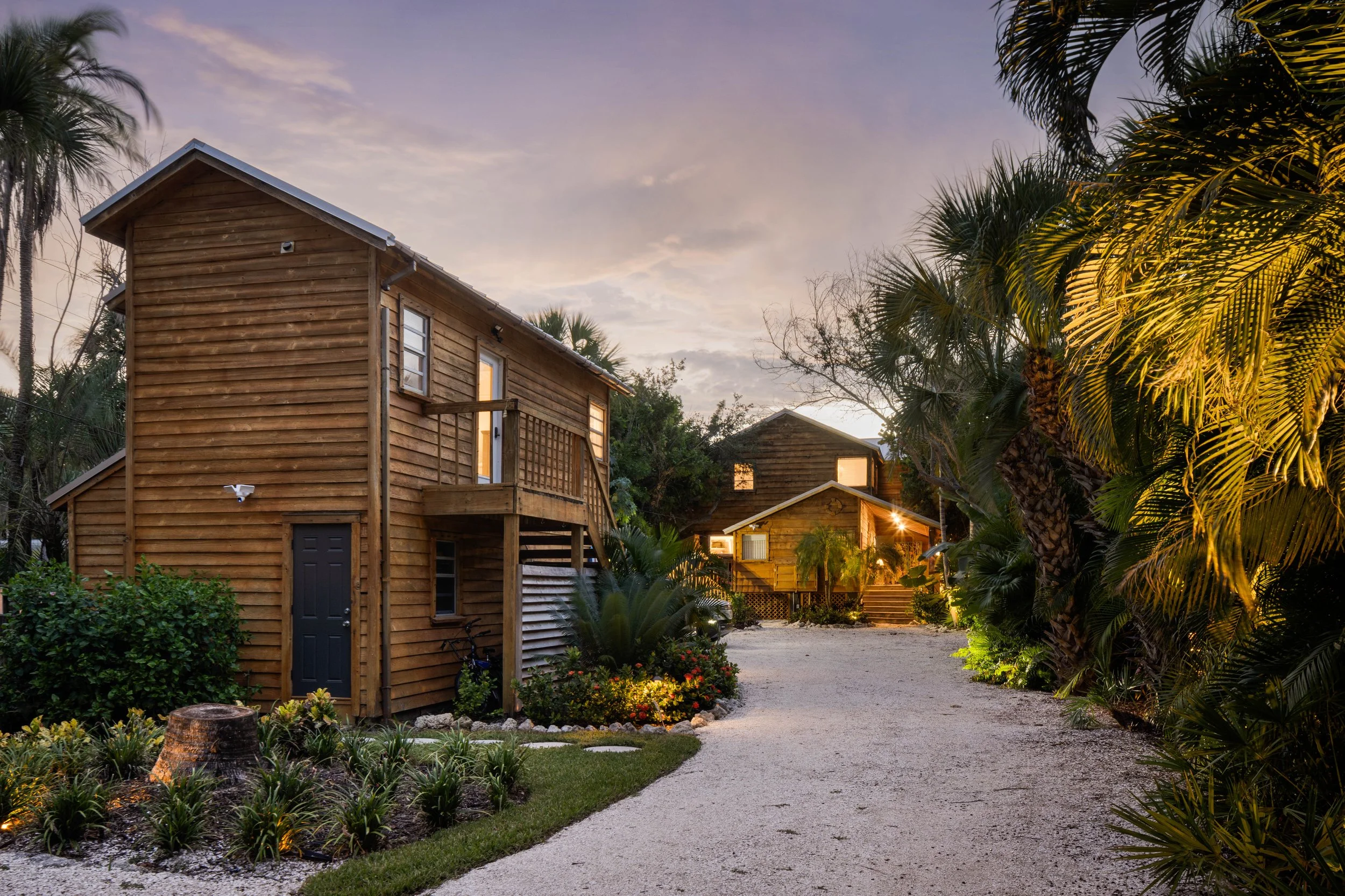 Wooden houses surrounded by tropical plants and palm trees with a gravel pathway at sunset.