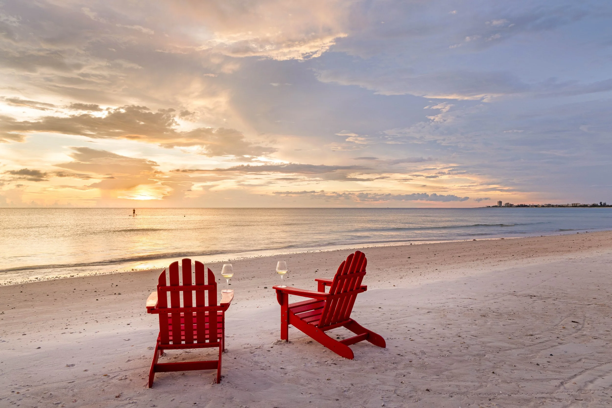 Two red Adirondack chairs facing the ocean on a sandy beach during sunset, with two glasses of white wine on a small table beside them and a person paddleboarding on the water in the distance. Original Beach House, Siesta Key, Florida