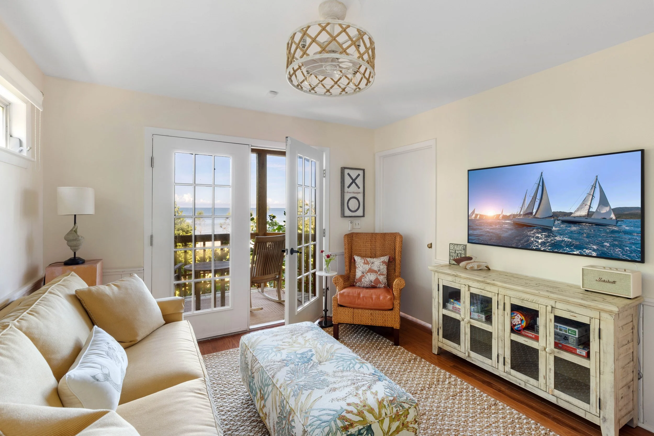A cozy living room with a beige couch, a patterned ottoman, an orange armchair, and a white distressed cabinet. French doors open to a balcony with a view of the ocean and sailboats. There is a wall-mounted flat-screen TV displaying sailboats, a sign that reads 'X O', and a woven ceiling light fixture.