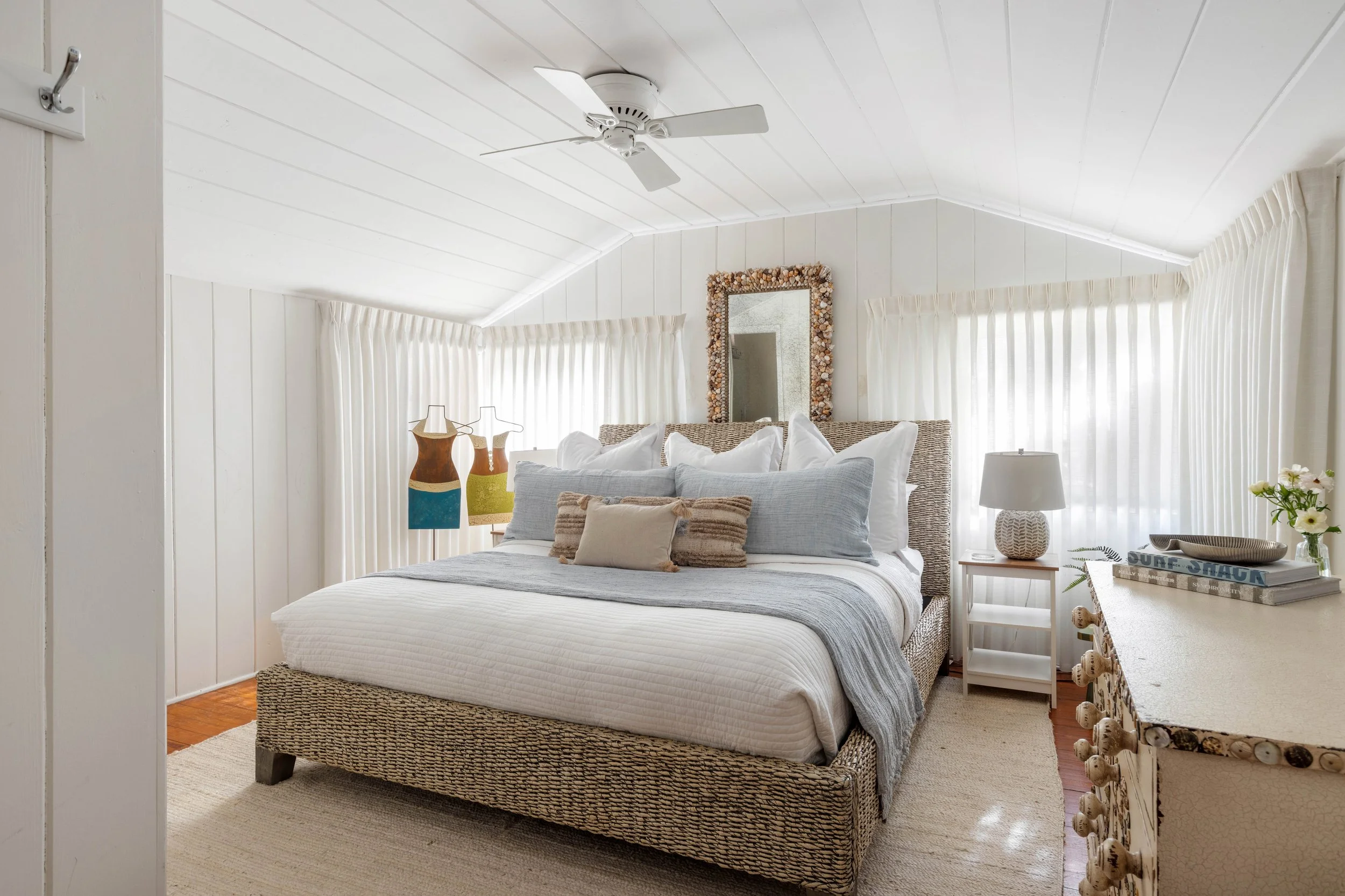 A cozy bedroom with a wicker bed featuring white bedding and decorative pillows. The walls and ceiling are painted white, with a ceiling fan. There are framed mirrors and colorful vases on the side tables, and sheer curtains cover the windows, lettin