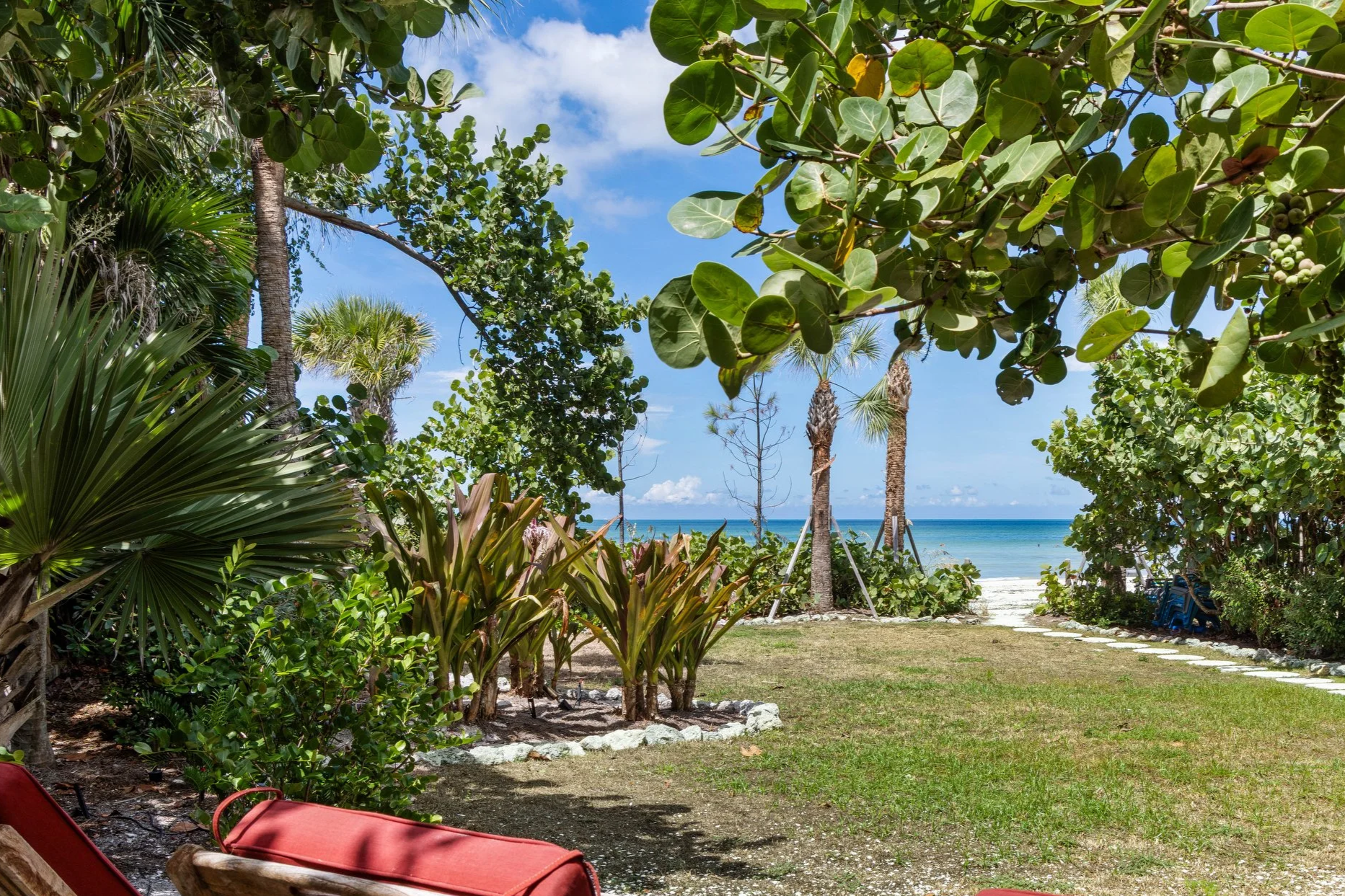 A tropical beach scene viewed through lush greenery, with palm trees and other tropical plants leading to a sandy beach and the ocean, under a partly cloudy sky. Original Beach House, Siesta Key, Florida