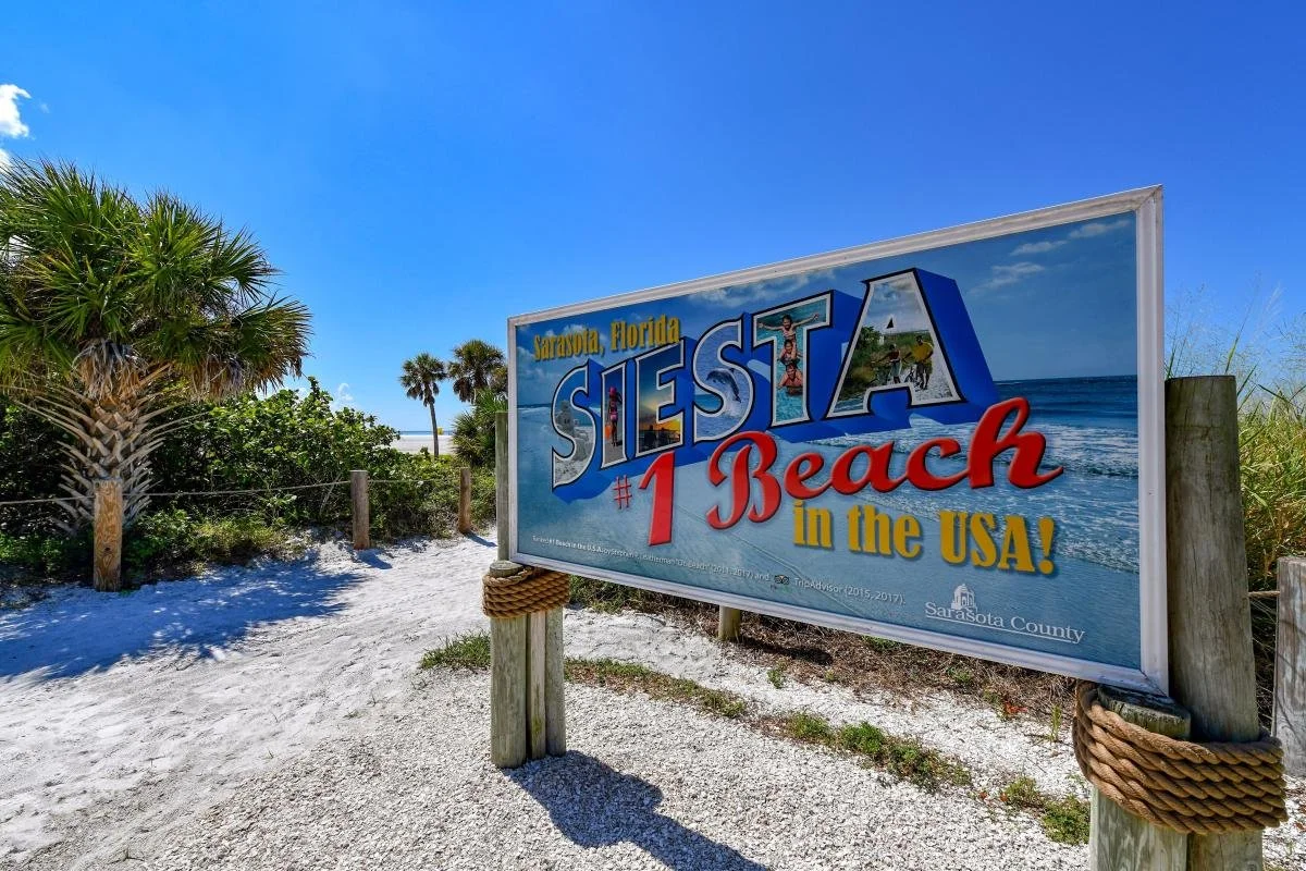 Sign for Siesta Beach in Sarasota, Florida, highlighting it as the number one beach in the USA, with a background of sandy pathway, palm trees, and a clear blue sky.