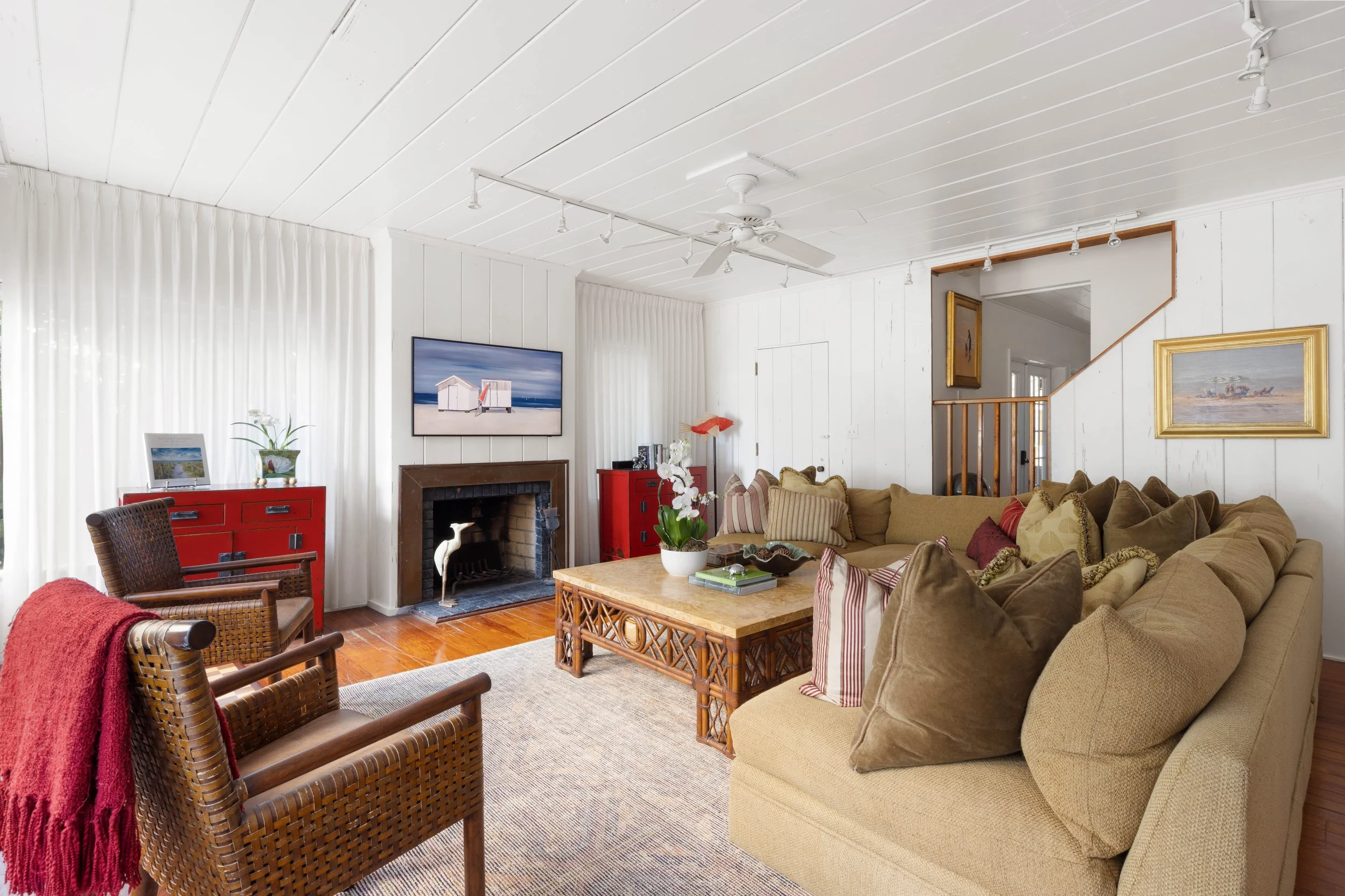 Living room with white paneled ceiling, beige sofa, wooden coffee table, brick fireplace, wall-mounted TV, plants, framed artwork, and red cabinets.