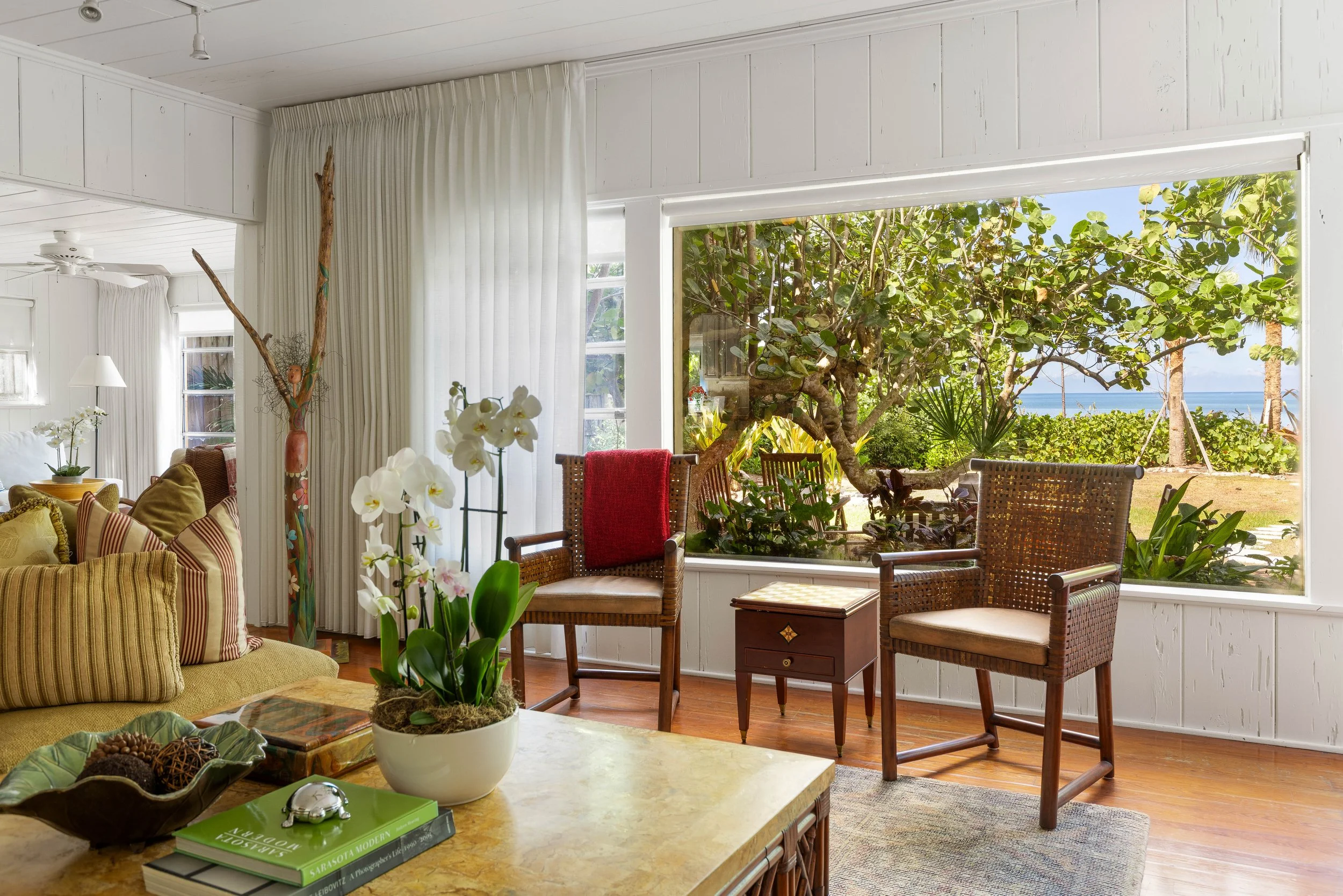 Living room with white walls and ceiling, beige sofa with striped cushions, potted orchids on coffee table, large window and sliding door opening to a garden with trees and ocean view.