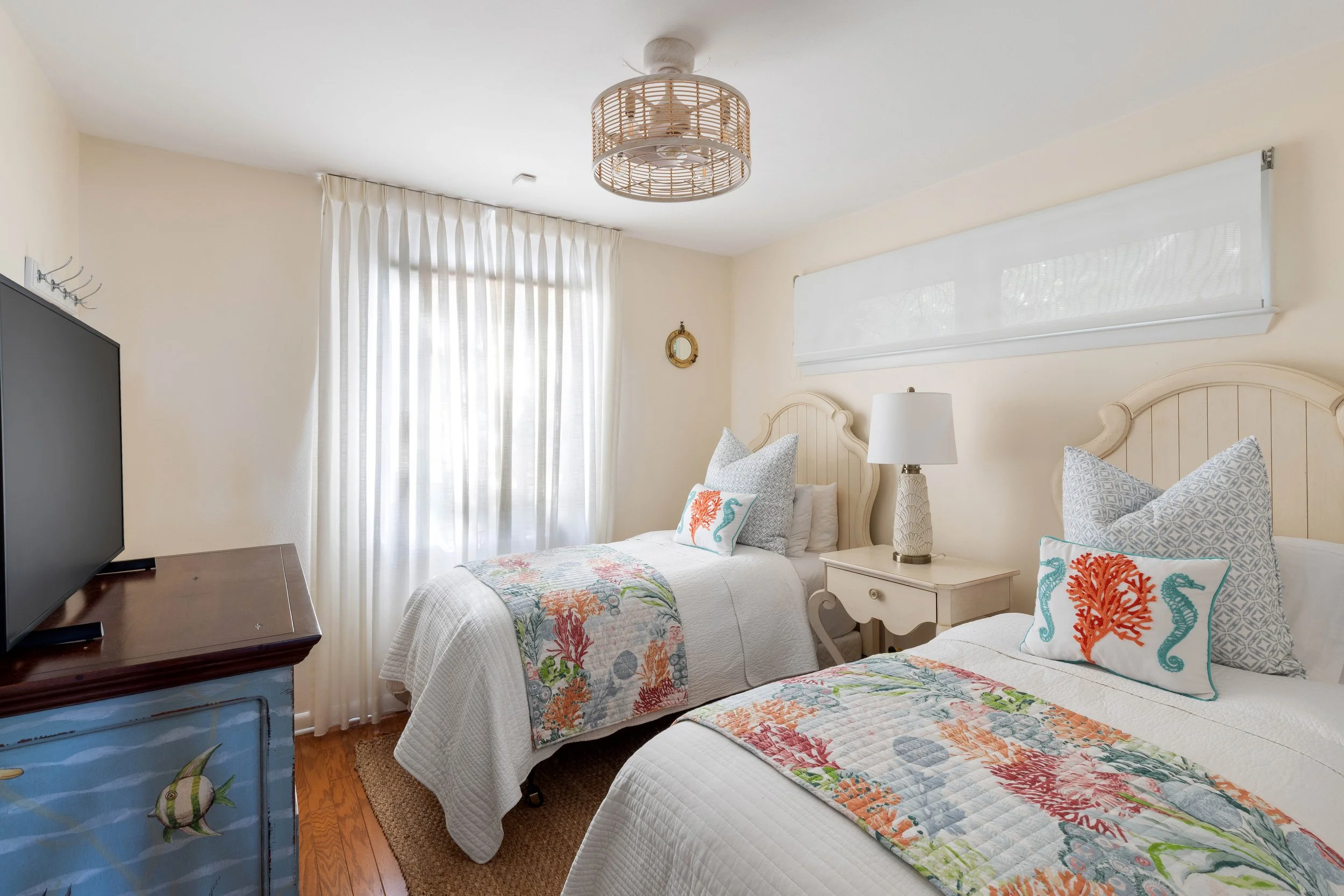 A bedroom with two twin beds, each with white bedding and coastal-themed pillows. There is a bedside table with a lamp between the beds, a window with sheer white curtains, a ceiling light fixture, and a dresser with a television.